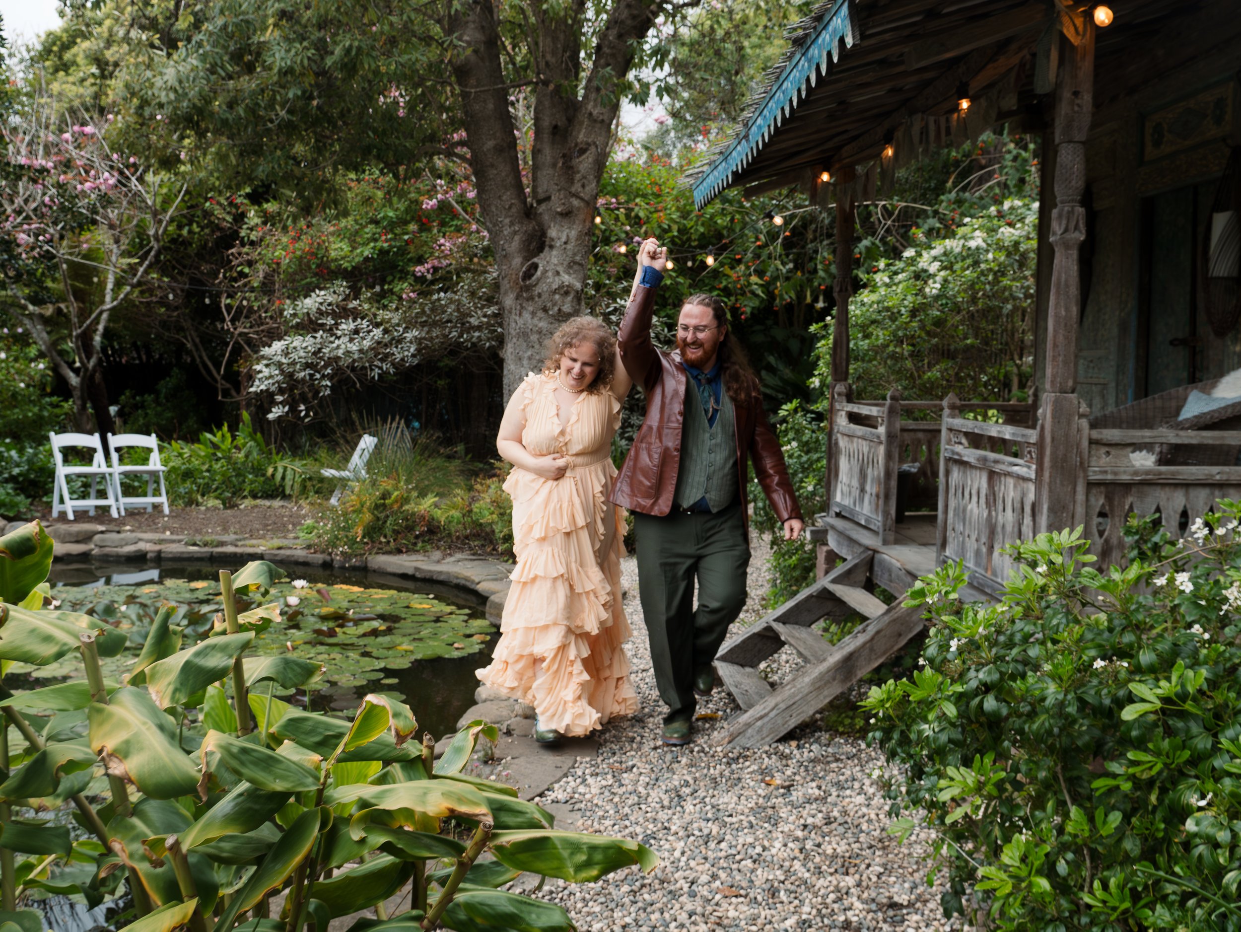 A couple walking hand-in-hand through a lush garden with a pond and lily pads, surrounded by trees and a wooden house with string lights.