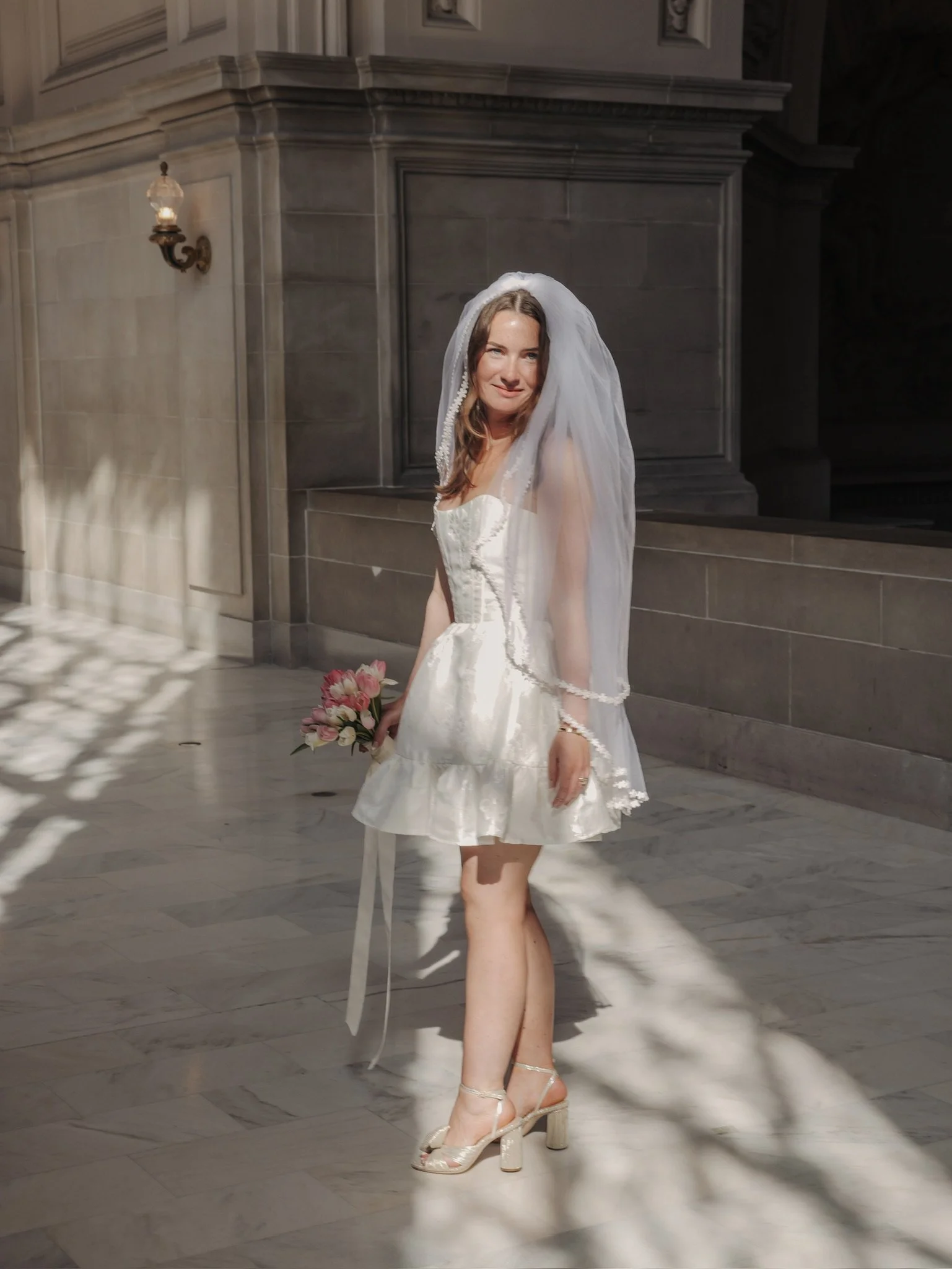 A young woman in a white wedding dress with a veil, holding a bouquet of pink flowers, standing inside San Francisco City Hall, wearing high-heeled sandals.