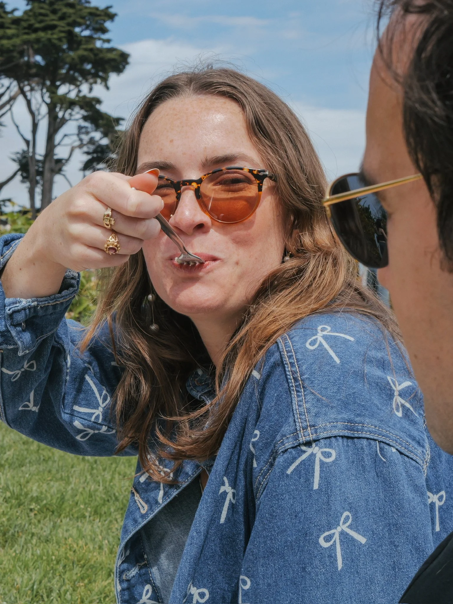 A woman wearing sunglasses and a denim jacket with white bow patterns is eating with a fork, with another person in sunglasses nearby, outdoors in a park with trees and grass.