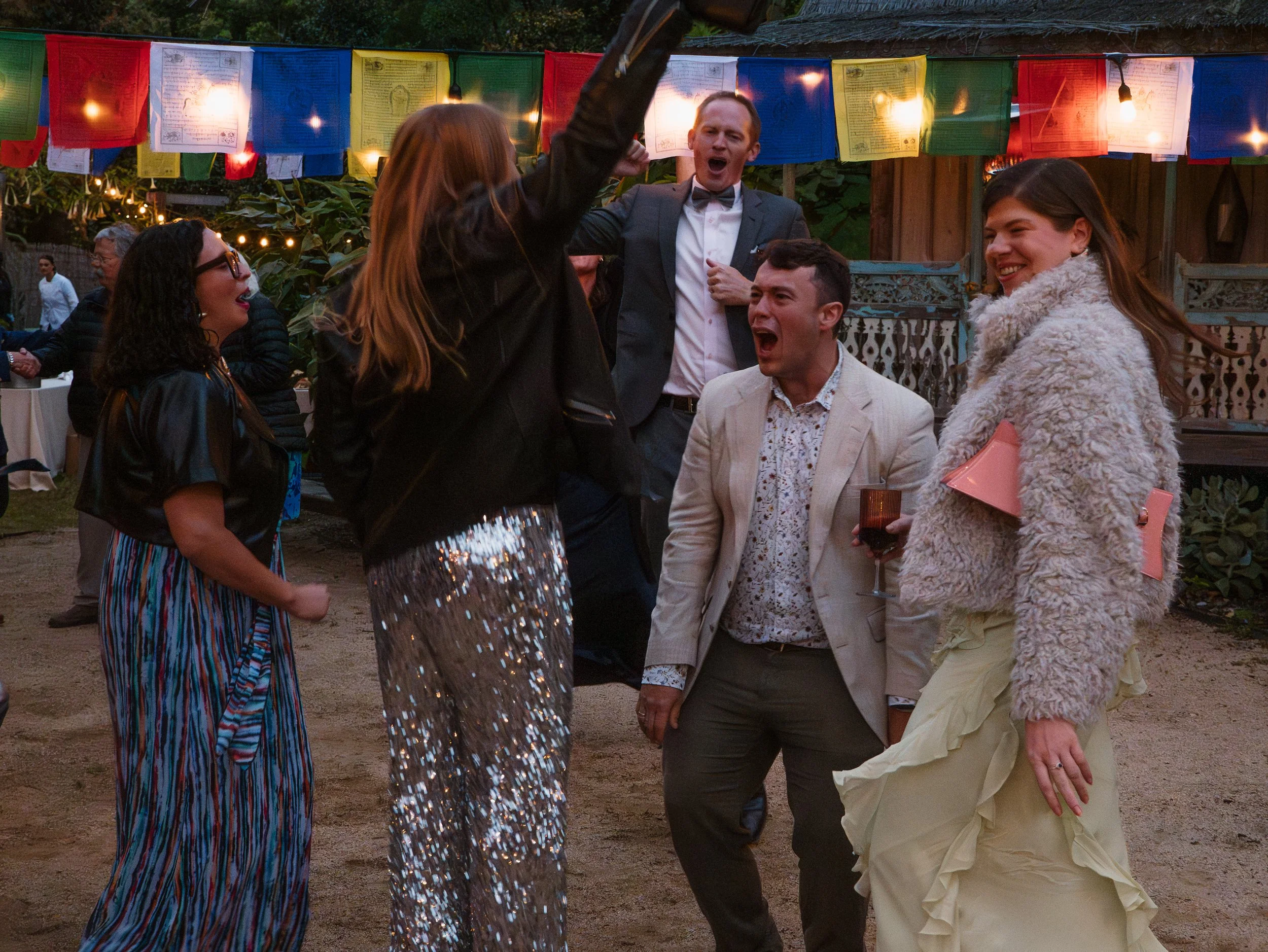 People dancing and socializing at an outdoor event decorated with colorful string flags and string lights during evening hours.