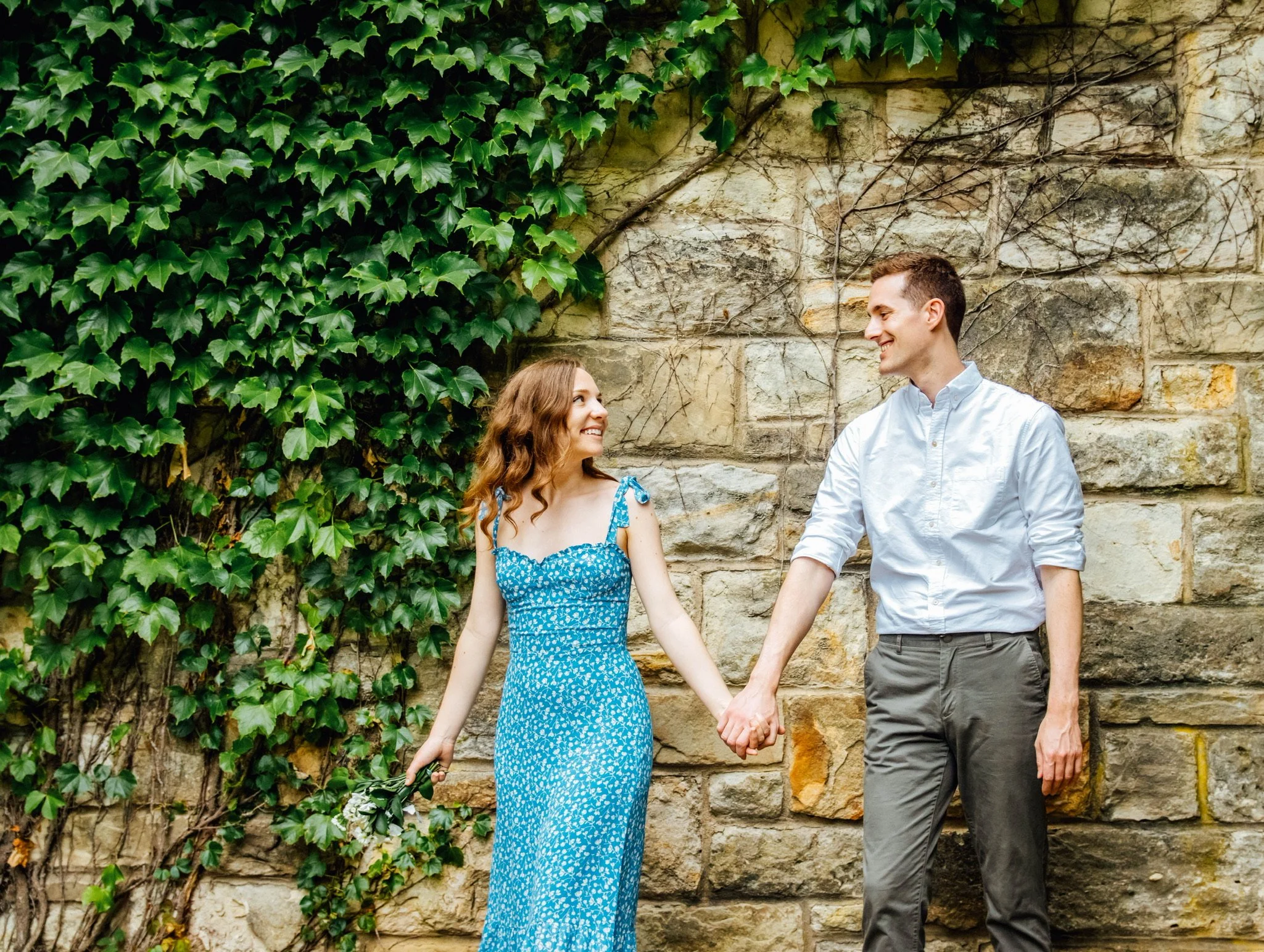 A young couple holding hands and smiling at each other outdoors, standing against a stone wall covered in green ivy.