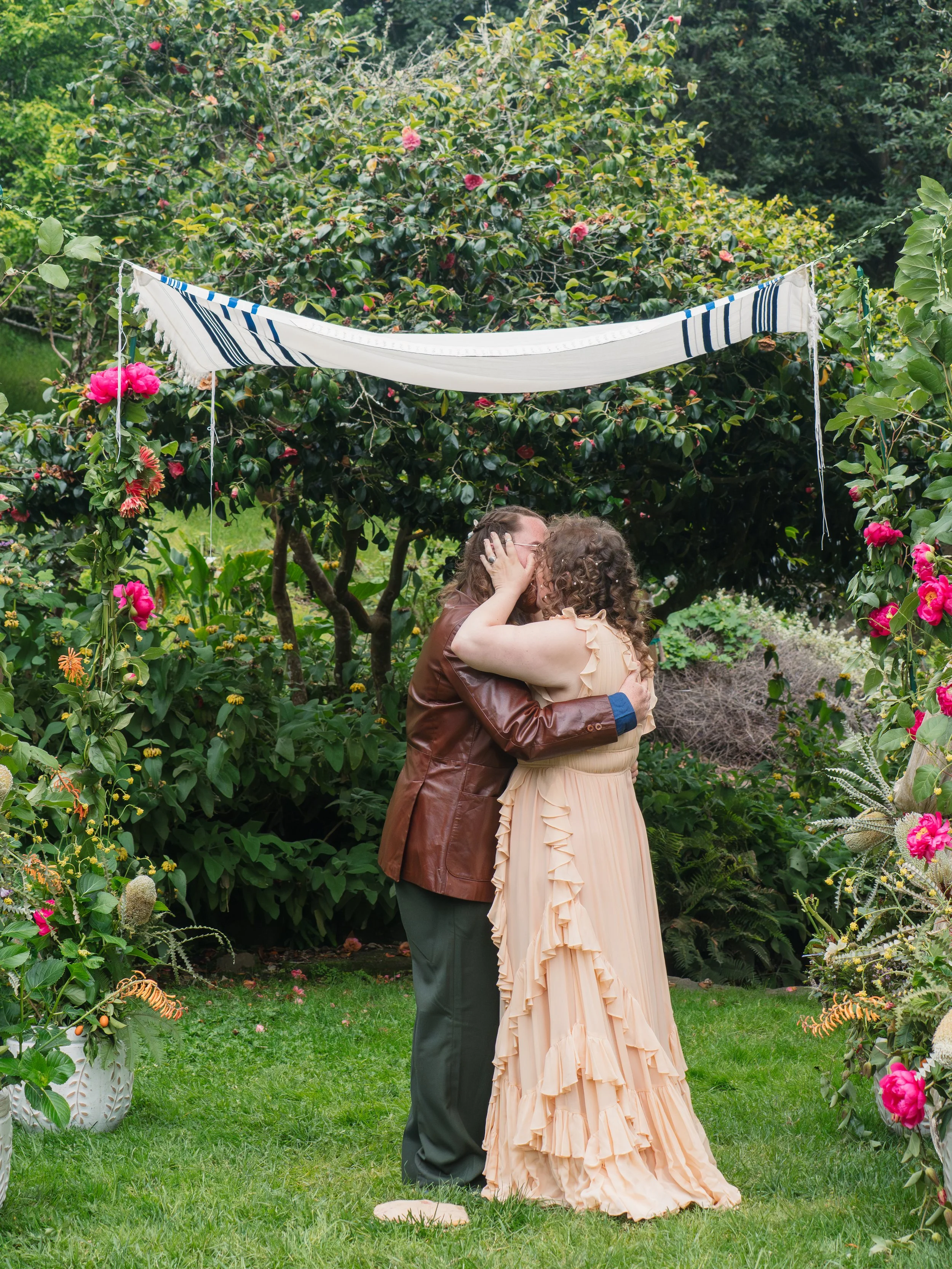 A couple shares a kiss in a lush garden setting, one wearing a beige ruffled dress and the other in a brown leather jacket and dark pants, surrounded by flowering bushes and greenery.
