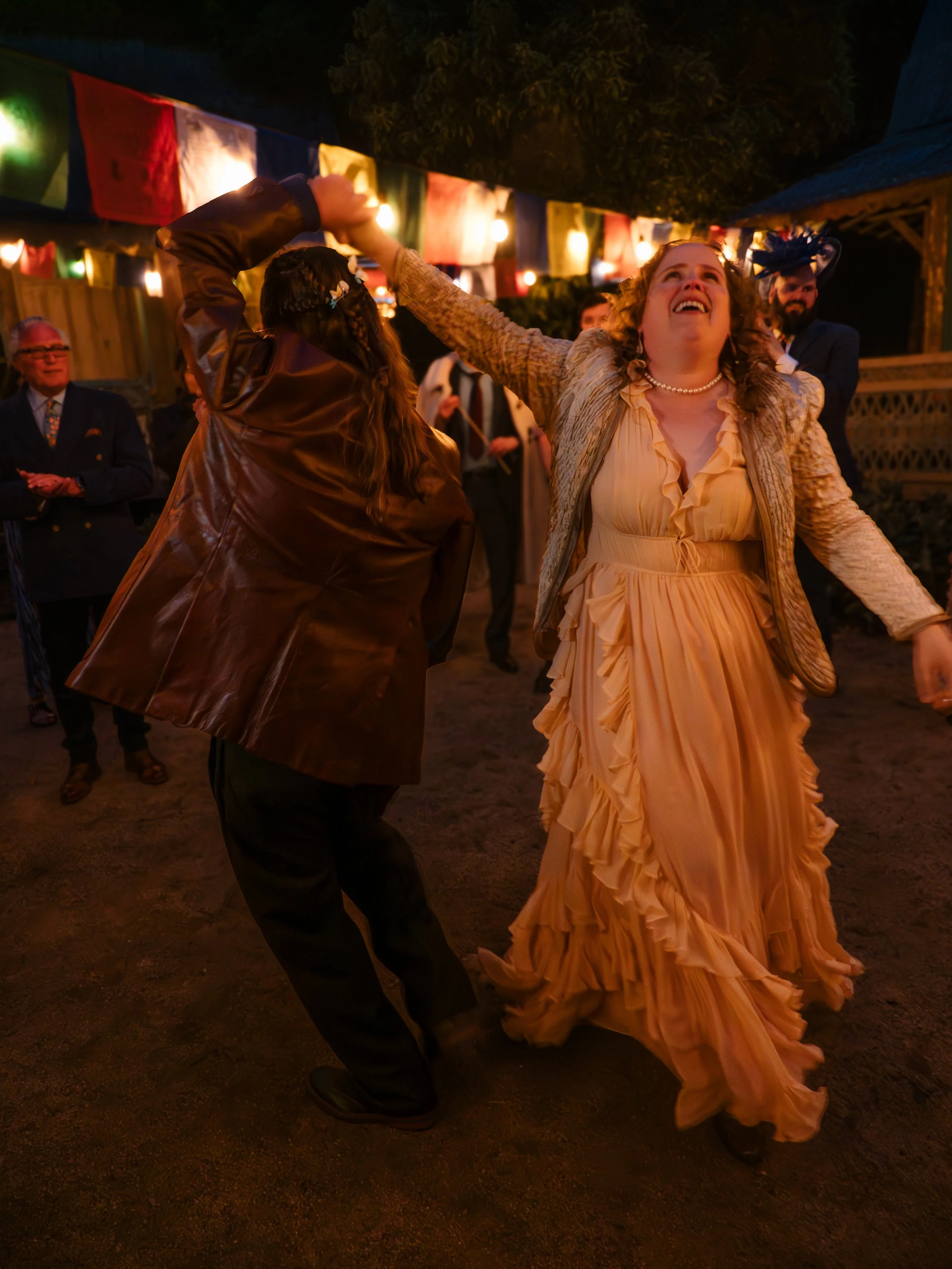 A woman in a peach dress dancing with a man in a brown jacket at an outdoor night celebration with colorful flags and string lights.