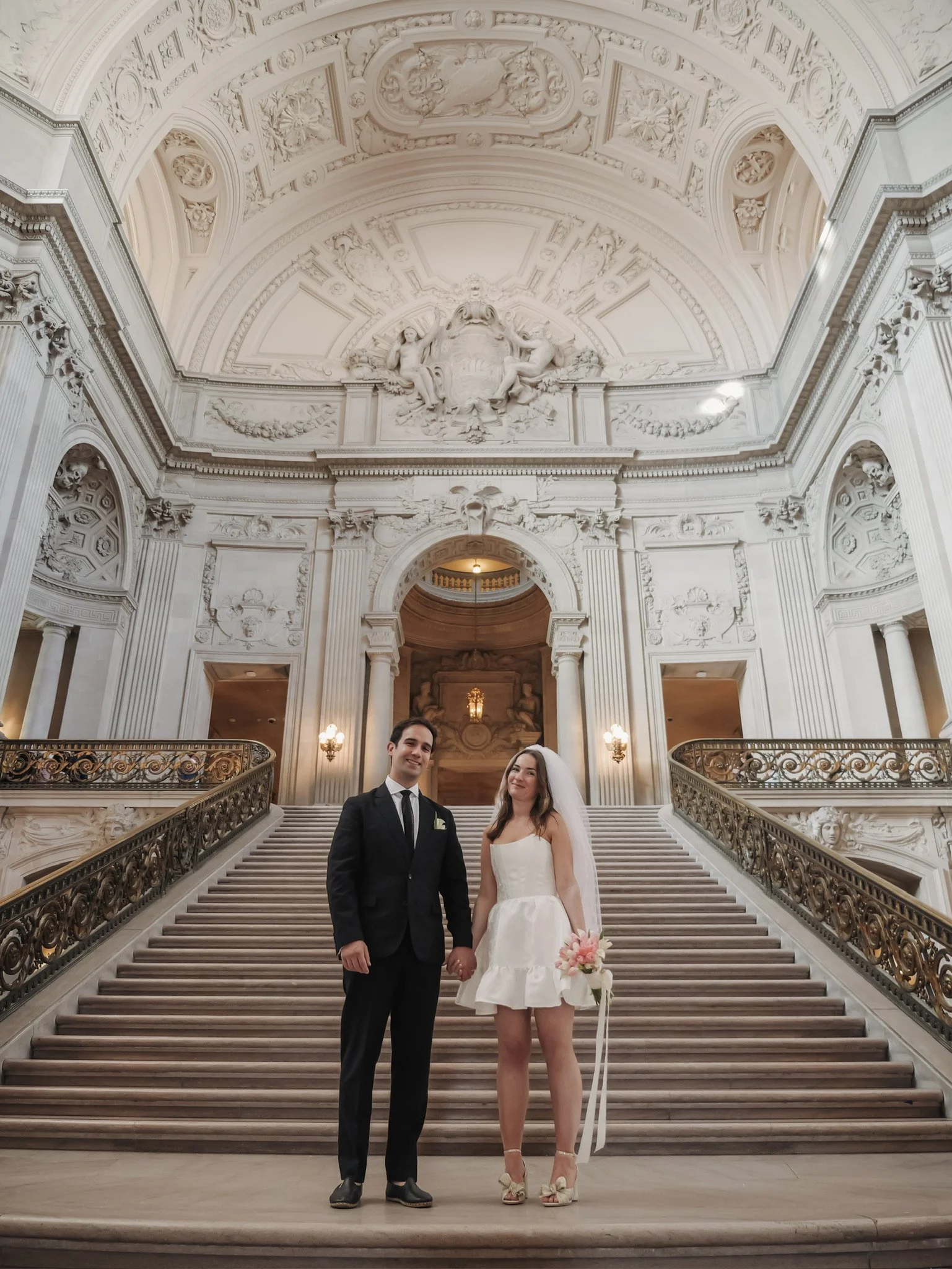 A bride and groom standing on a grand staircase in San Francisco City Hall with ornate white ceilings and decorative sculptures, dressed in wedding attire, holding hands and smiling.
