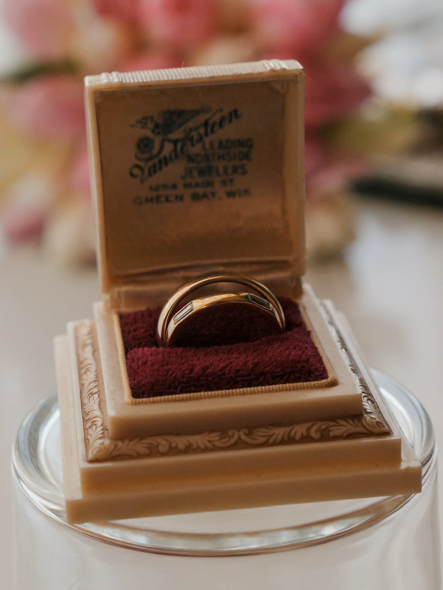 A small, open jewelry box with ornate detailing on the edges, containing a single shiny ring inside. The box is lined with red velvet and is placed on a clear glass surface.