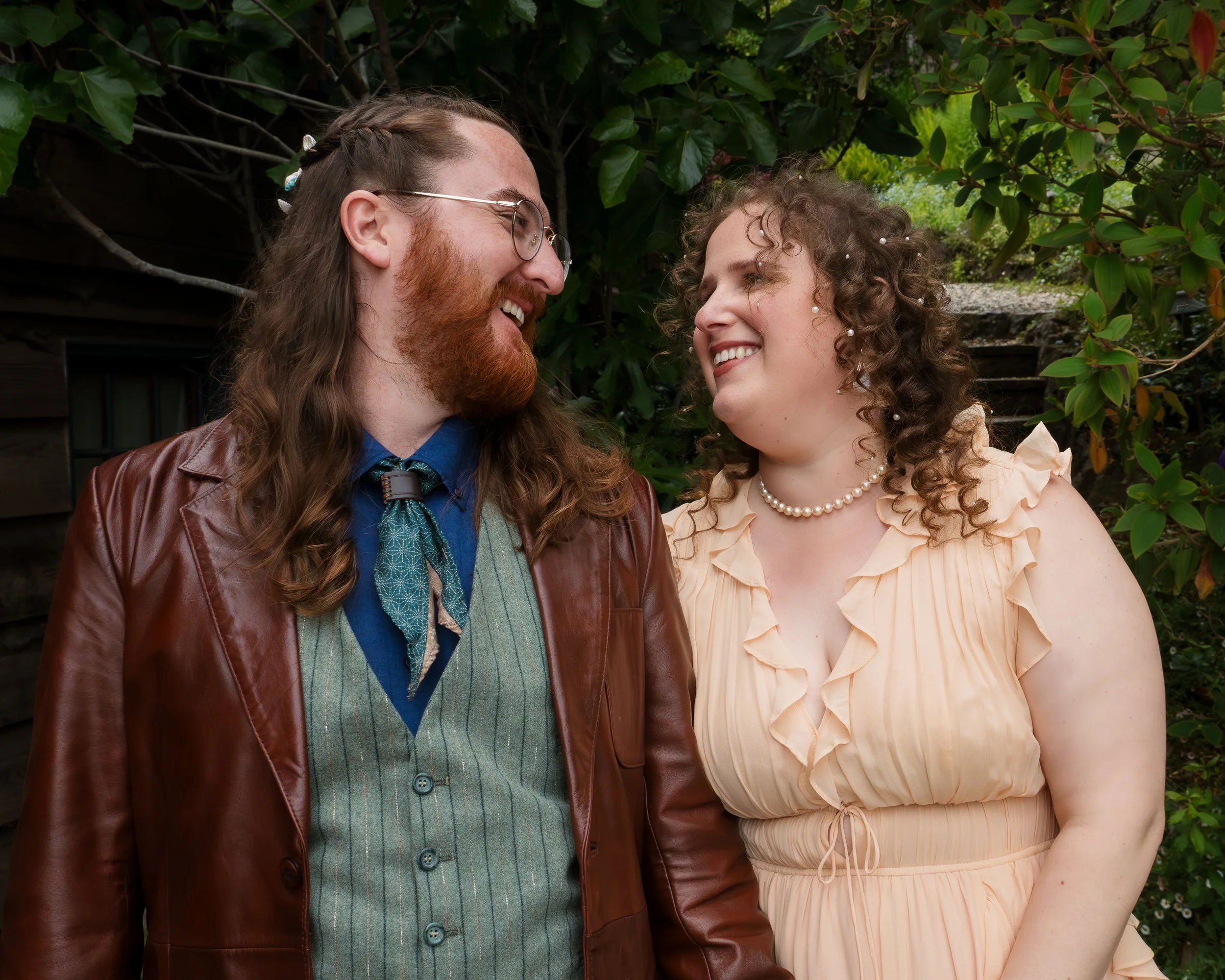 A man and woman standing close together outdoors, smiling and looking at each other, surrounded by green plants.