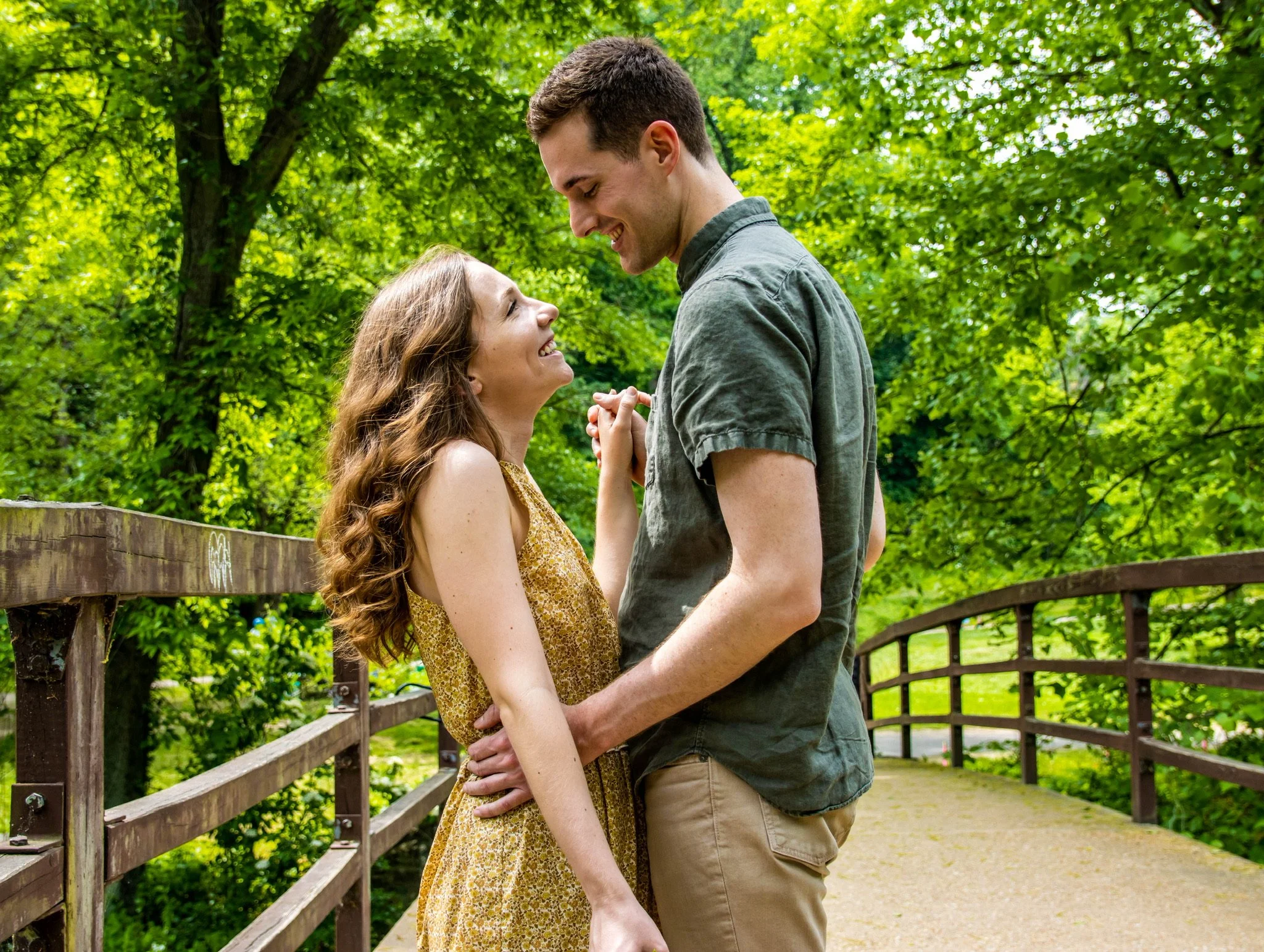 A young couple standing on a wooden bridge surrounded by lush green trees, smiling at each other and holding hands.