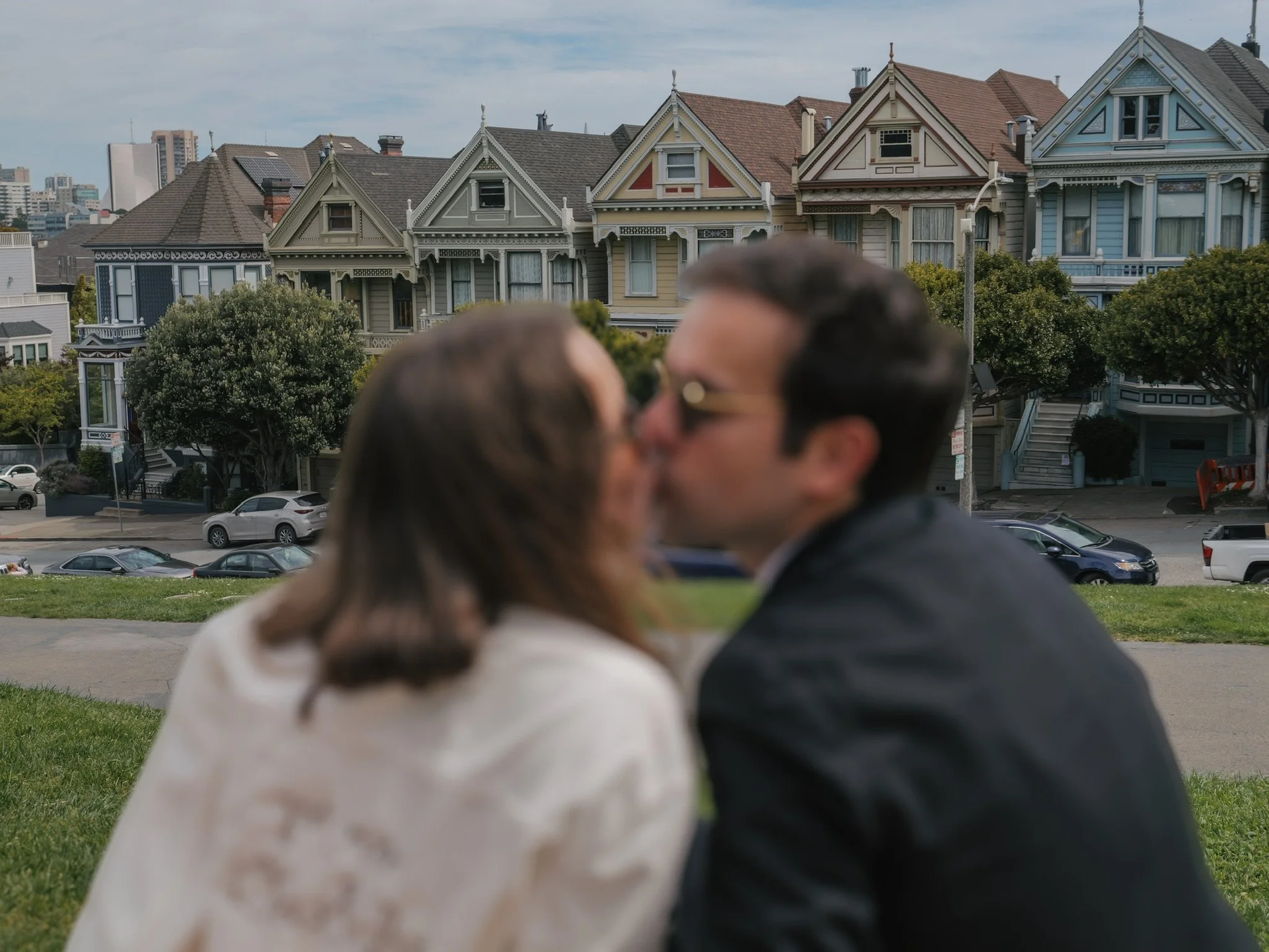A couple kissing outdoors with Victorian-style houses and trees in the background.