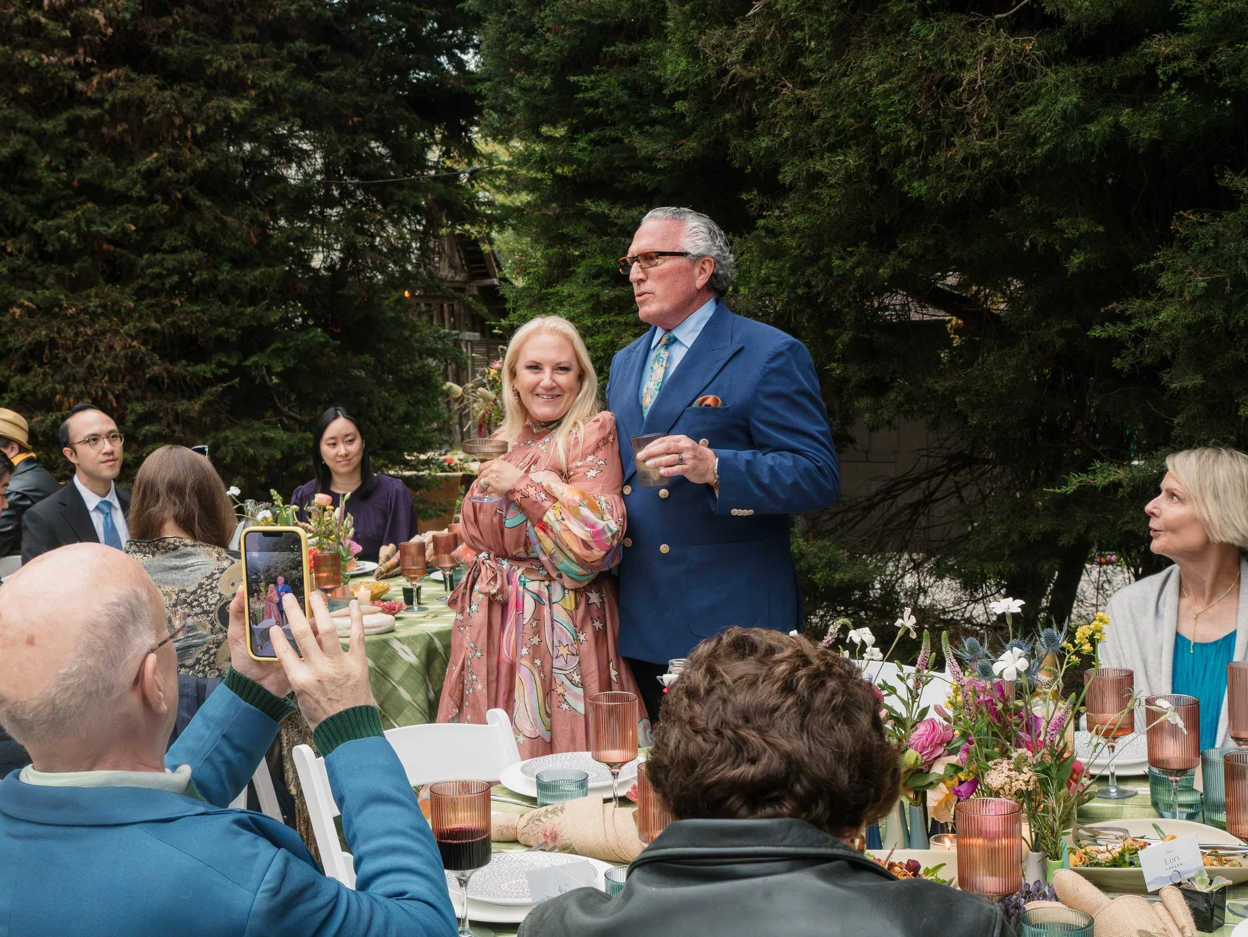 A group of people gathered outdoors for a celebration, with a woman in a pink dress and a man in a blue suit standing near a table with food and drinks. There are trees in the background and someone is taking a photo of the scene.