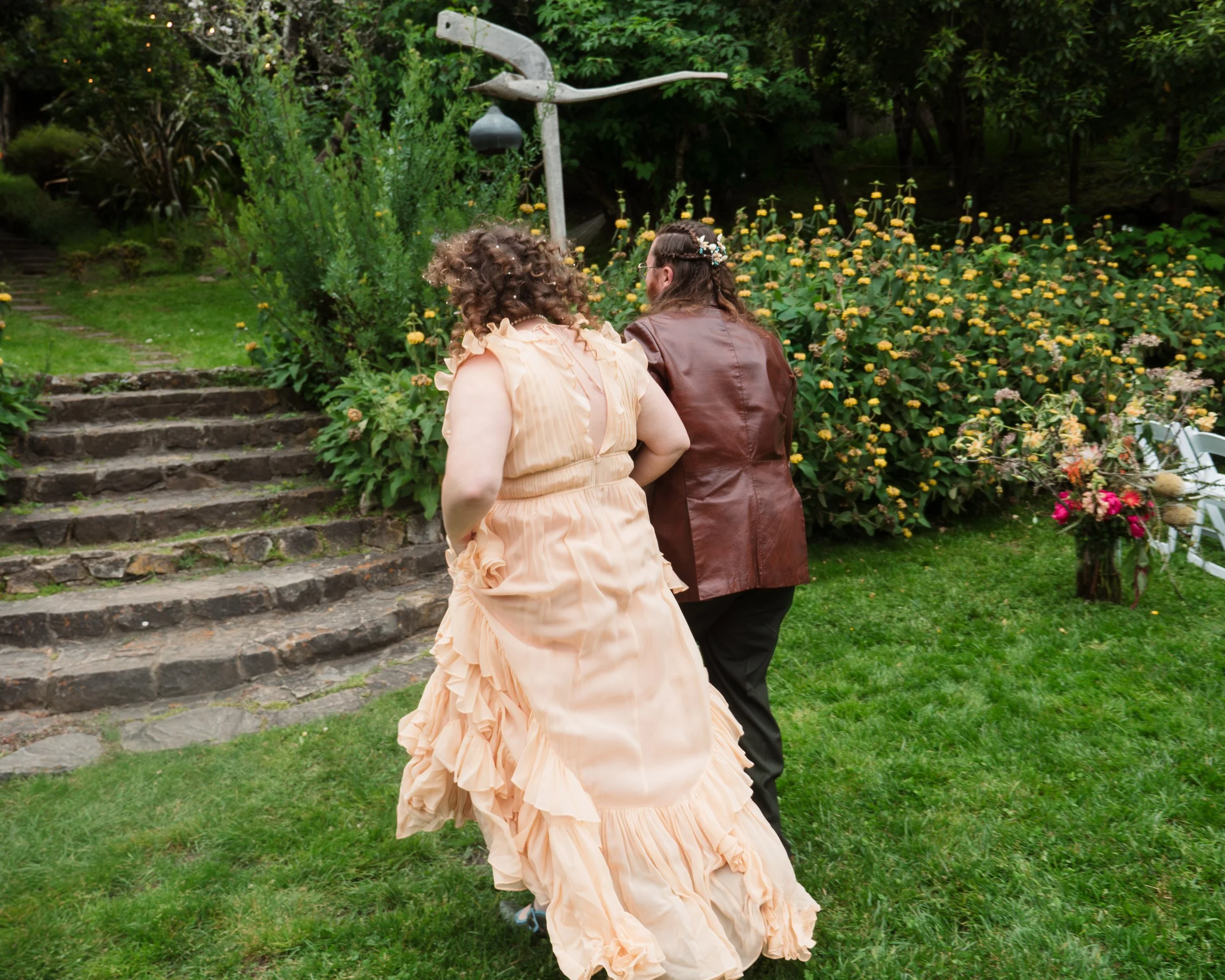 A couple walks up stone steps in a lush garden with flowers and trees.