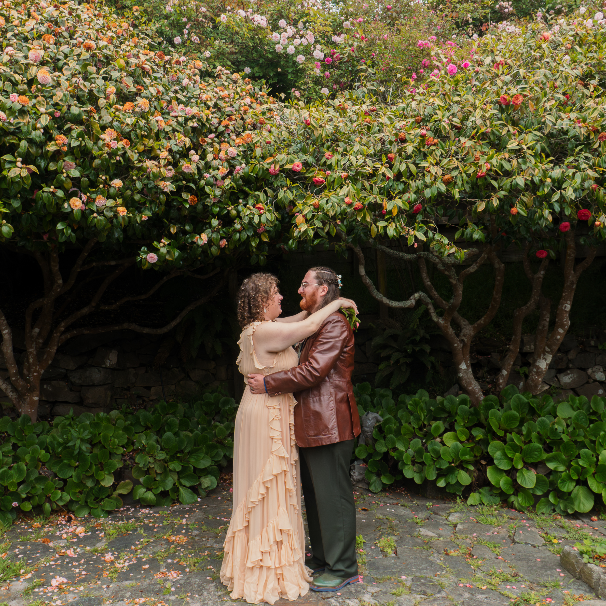 A couple embraces in front of a lush green bush with pink and cream flowers, outdoors on a stone pathway with fallen flower petals.