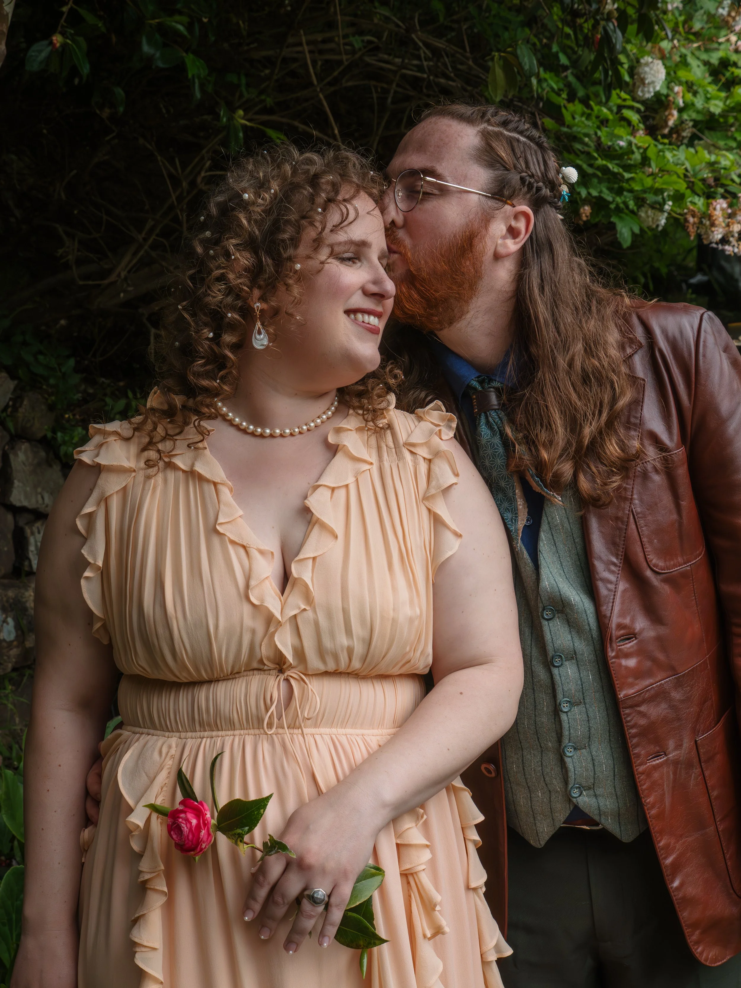 A couple dressed in vintage clothing, with the man kissing the woman's forehead in a garden setting. The woman has curly hair with small pearls, wearing earrings, a peach-colored dress with ruffles, a pearl necklace, and holding a pink rose. The man 