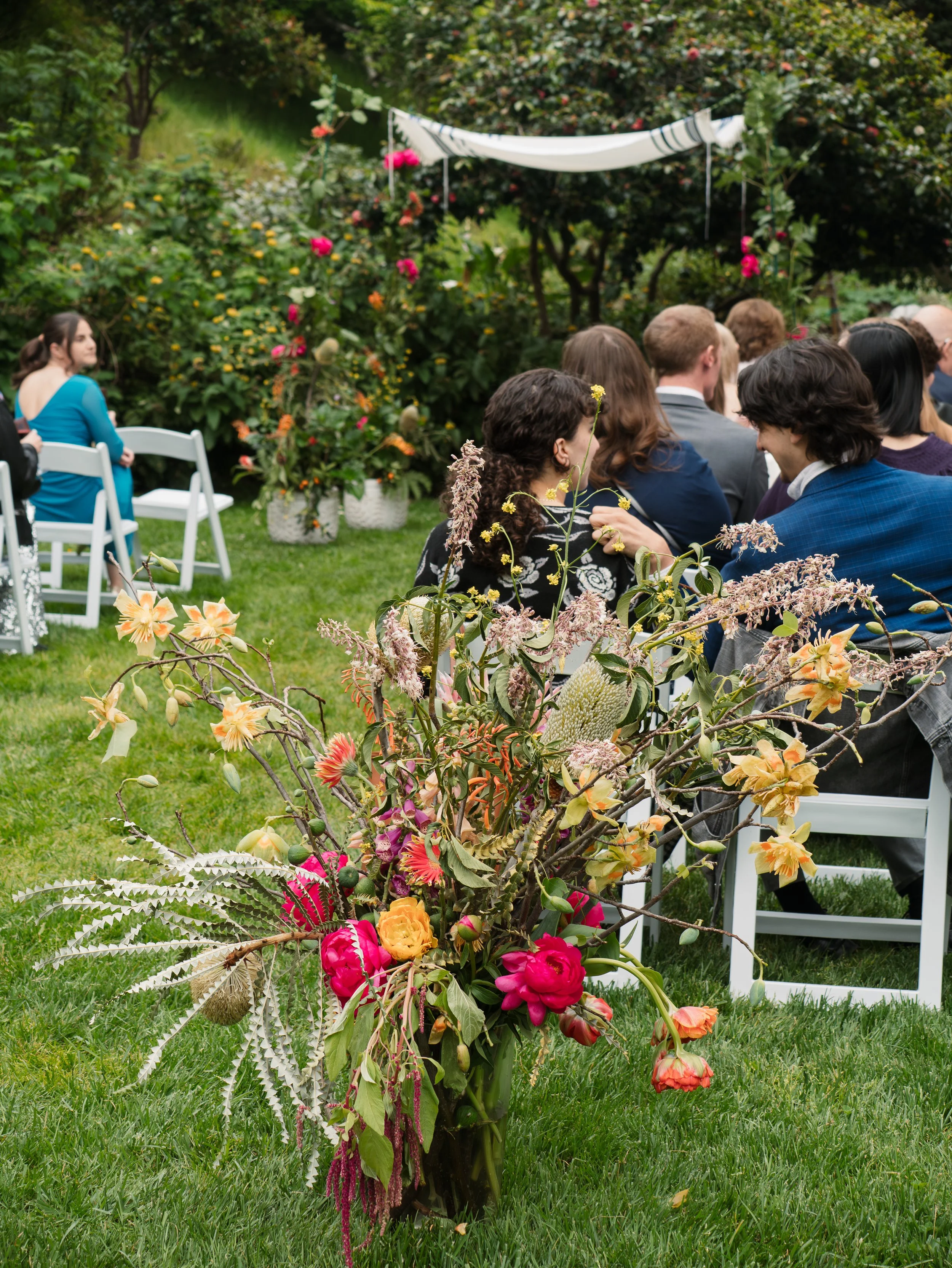 A large bouquet of colorful flowers on a grassy lawn in front of a group of people sitting on white chairs, with a lush green garden and trees in the background.