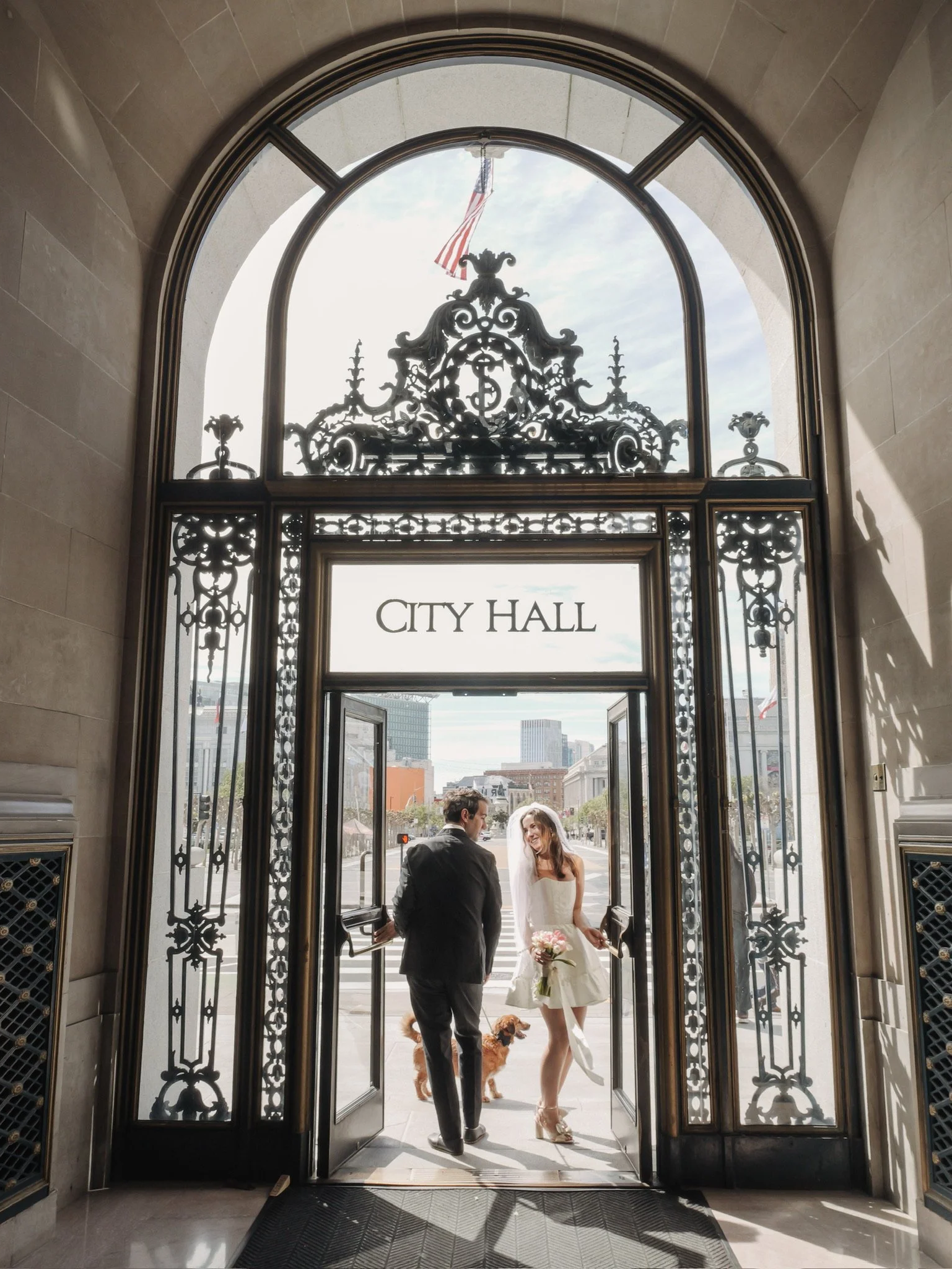 A bride and groom standing outside a city hall building, holding hands, with two dogs. The city hall has ornate black iron gates and an arched window with a flag flying outside.