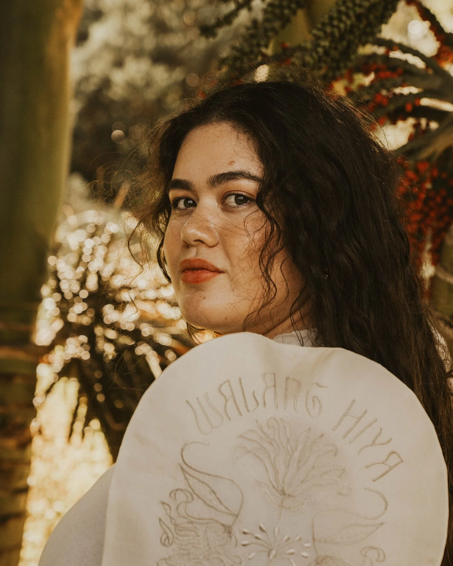 Close-up of a young woman with dark curly hair and light skin looking over her shoulder, with a background of trees with white and red leaves and blurred lights.