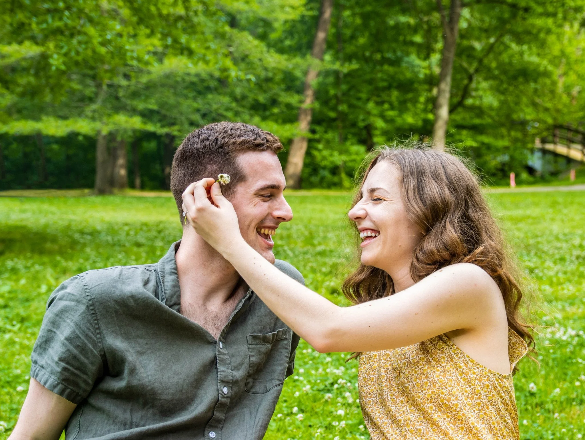 A young man and woman outdoors in a park, smiling and touching foreheads, with the woman holding a small white flower near his face.