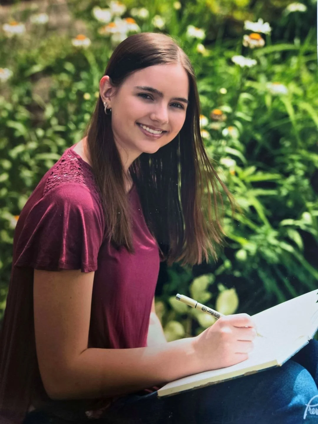 Image of Kaylee Pruitt with long brown hair and earrings, sitting outdoors in a garden with green foliage and flowers, smiling and writing in a notebook.