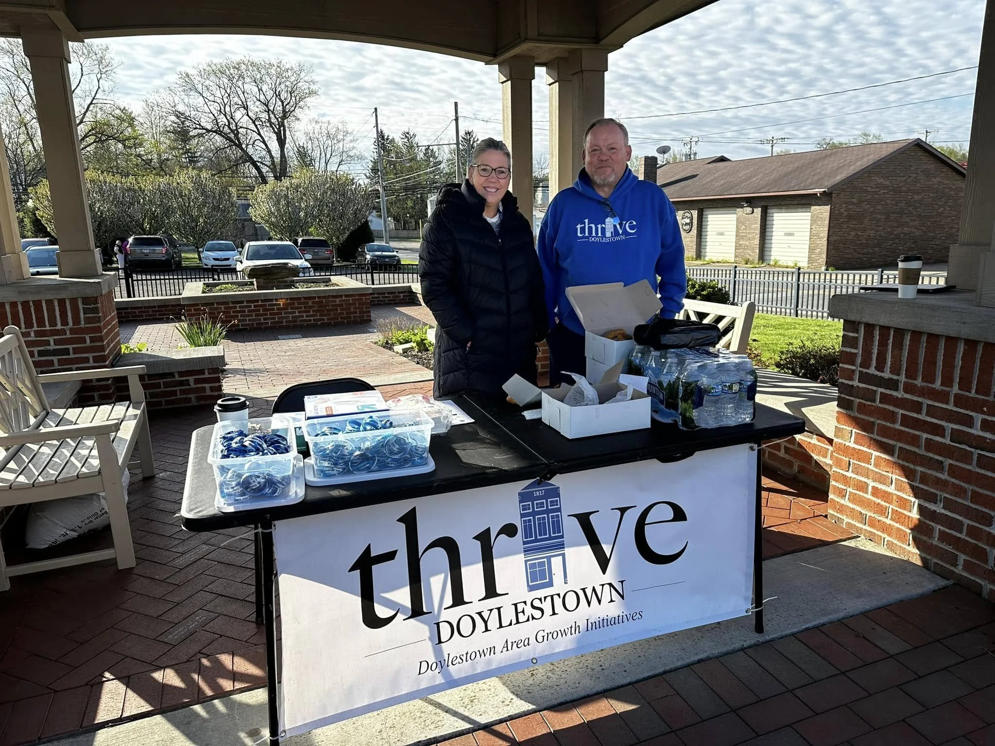 Two people standing behind a table with Thrive Doylestown signage, outdoor setting with parked cars and trees in the background
