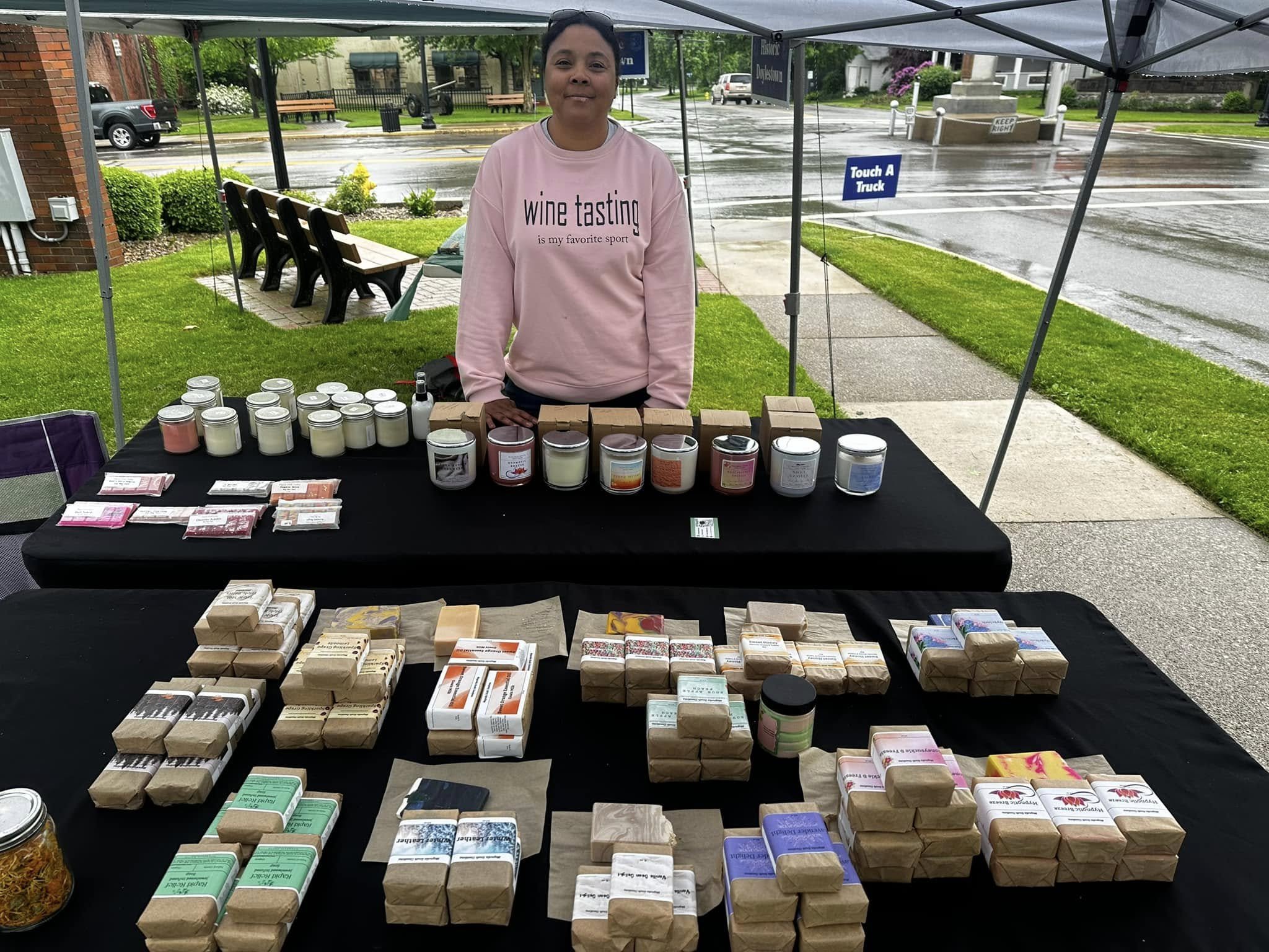 Woman standing at an outdoor booth with various soap and skincare products on display during a rainy day.
