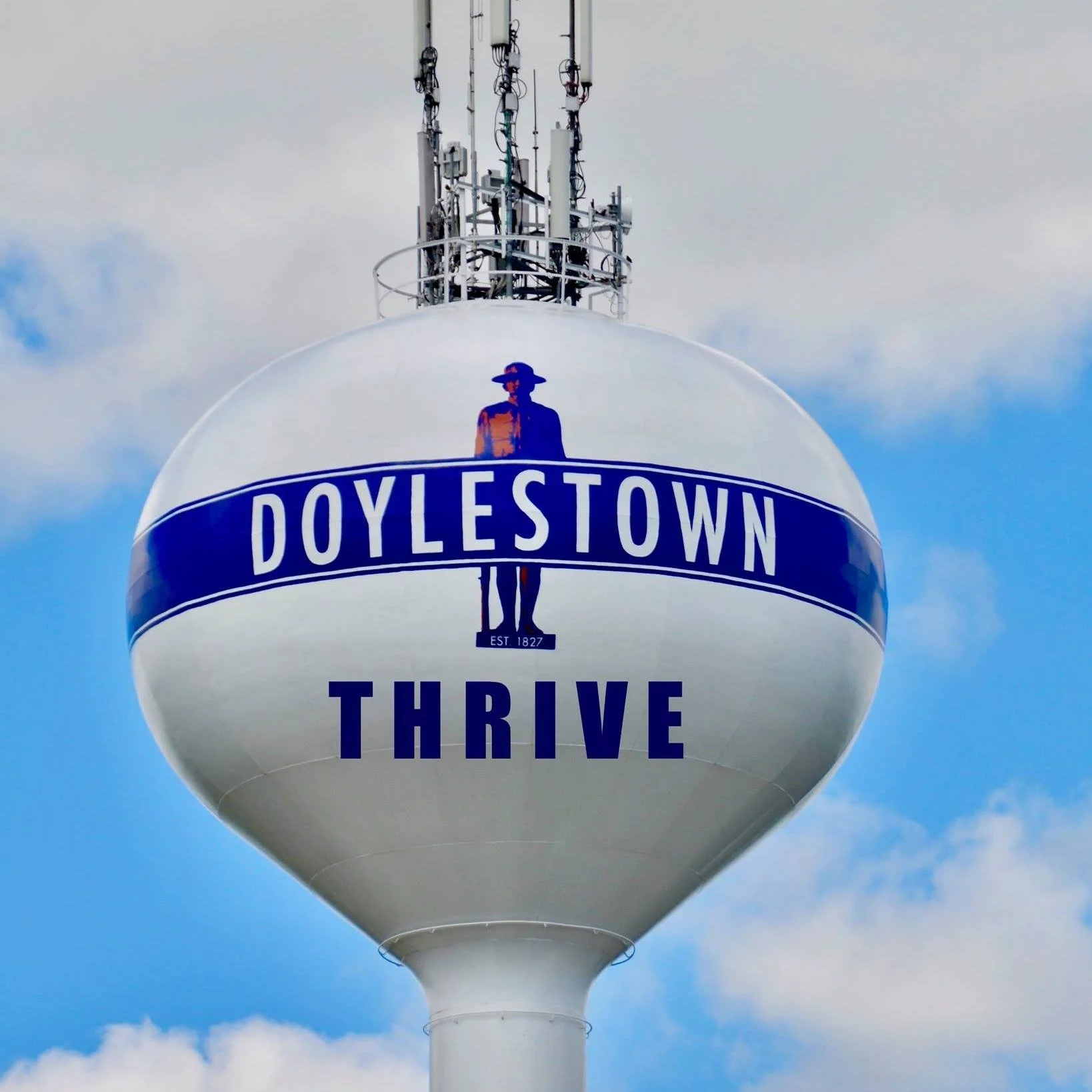 A water tower with a spherical top displays the text "Doylestown" and the phrase "Thrive" beneath a logo of a person wearing a hat, standing on a small platform, with a cloudy sky in the background.