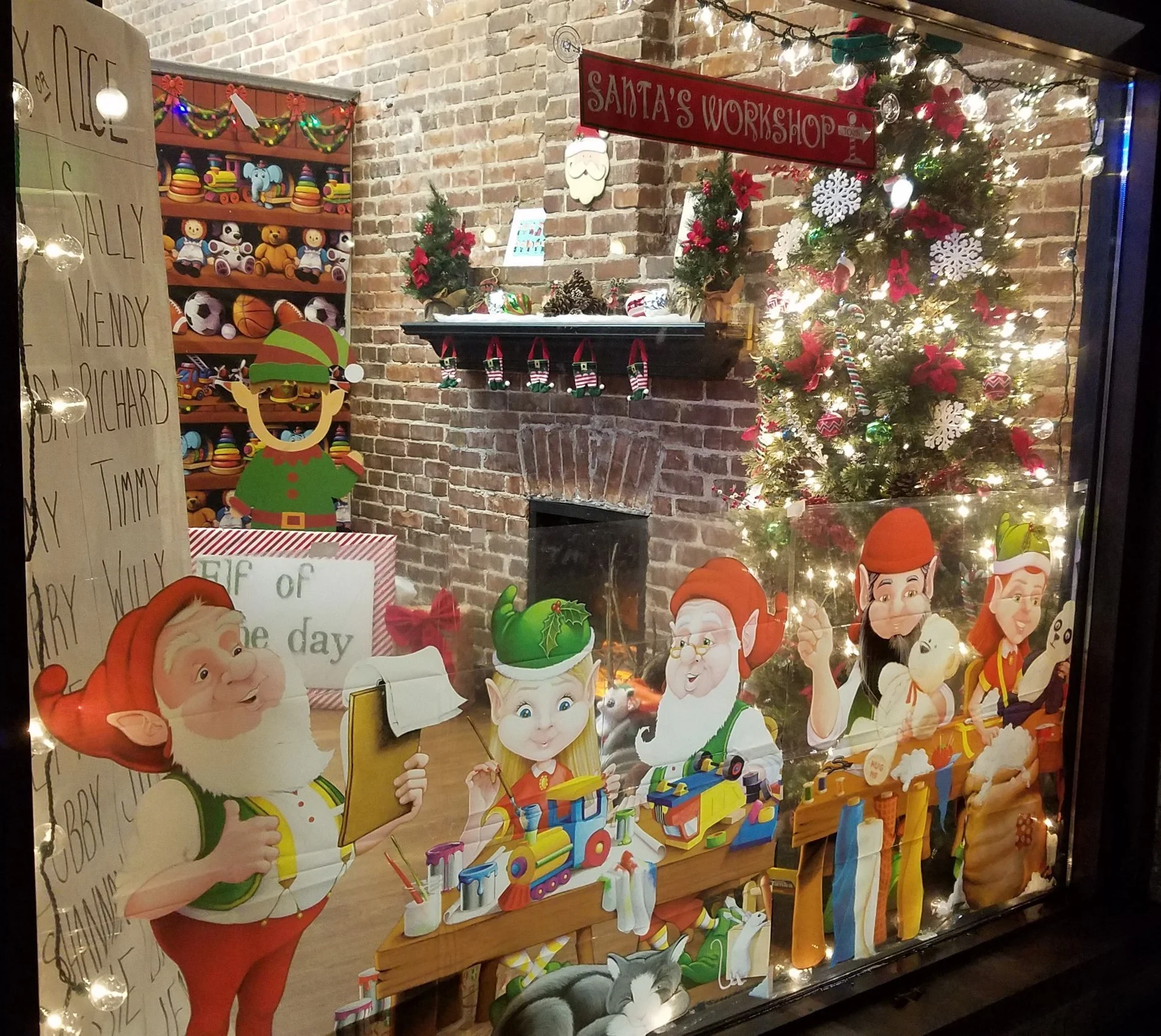 Christmas shop window display featuring animated characters, a decorated Christmas tree with lights, and festive decorations on a brick wall with a sign reading 'Santa's Workshop'.