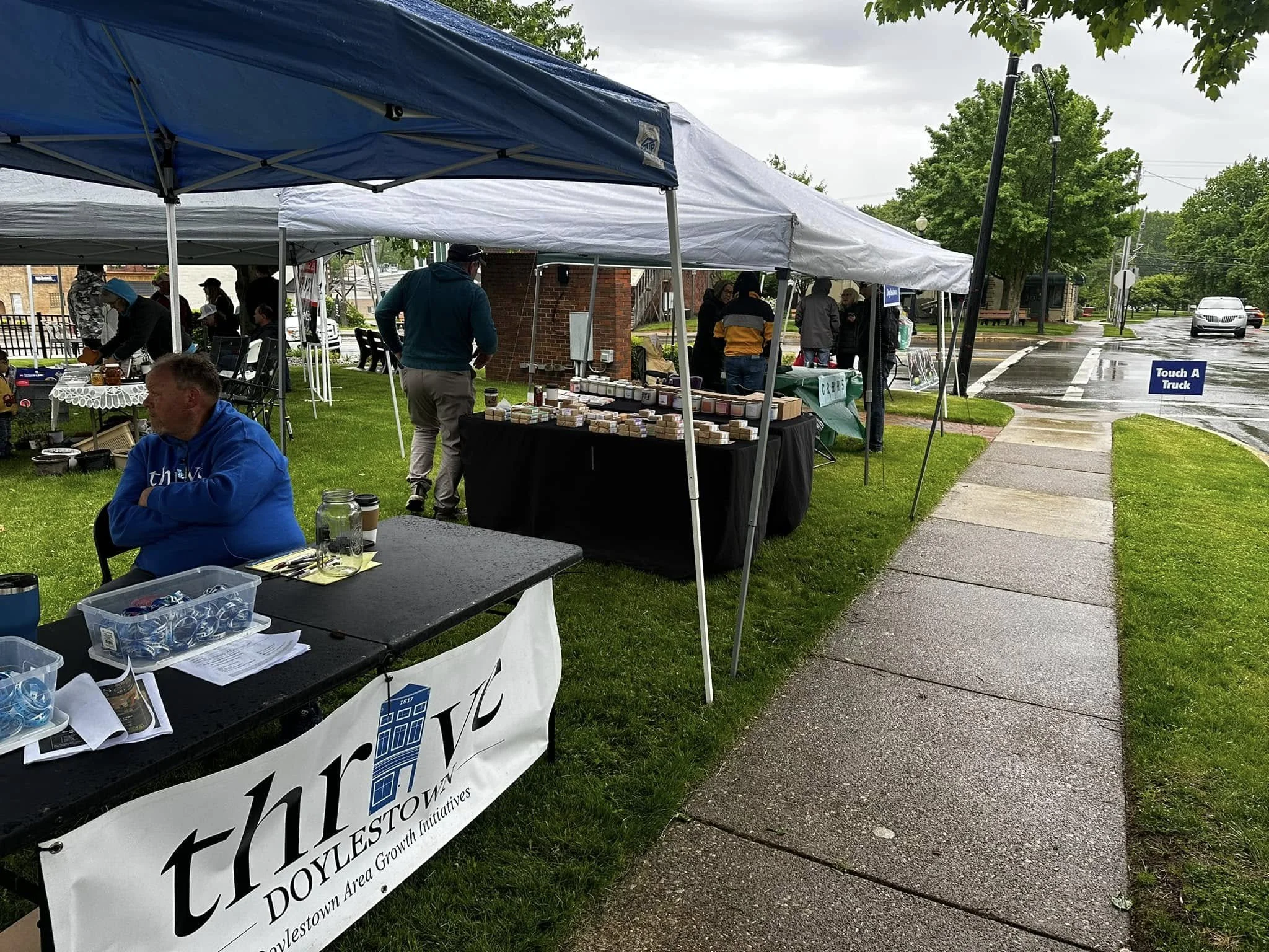 Outdoor market with multiple white and blue tents on a wet sidewalk, people browsing stalls, a man in a teal jacket, and a sign that reads 'Touch A Truck' on the street.