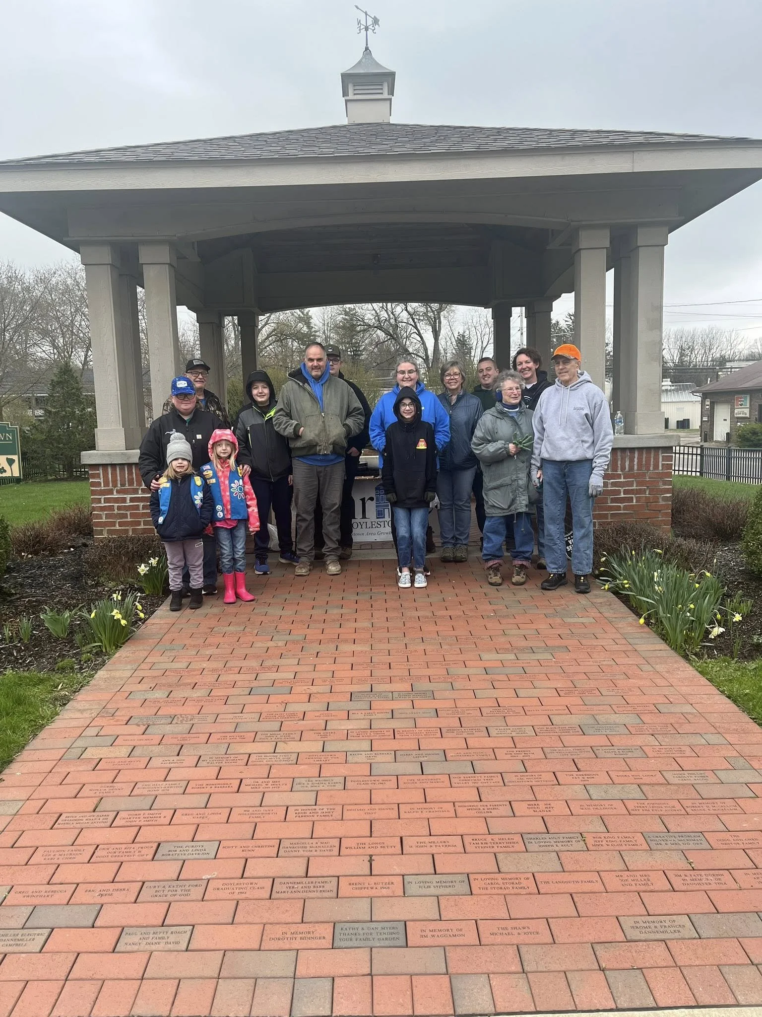 Group of people standing on a brick walkway in front of a memorial wall with engraved names, under a pavilion in an outdoor setting with trees and buildings in the background.