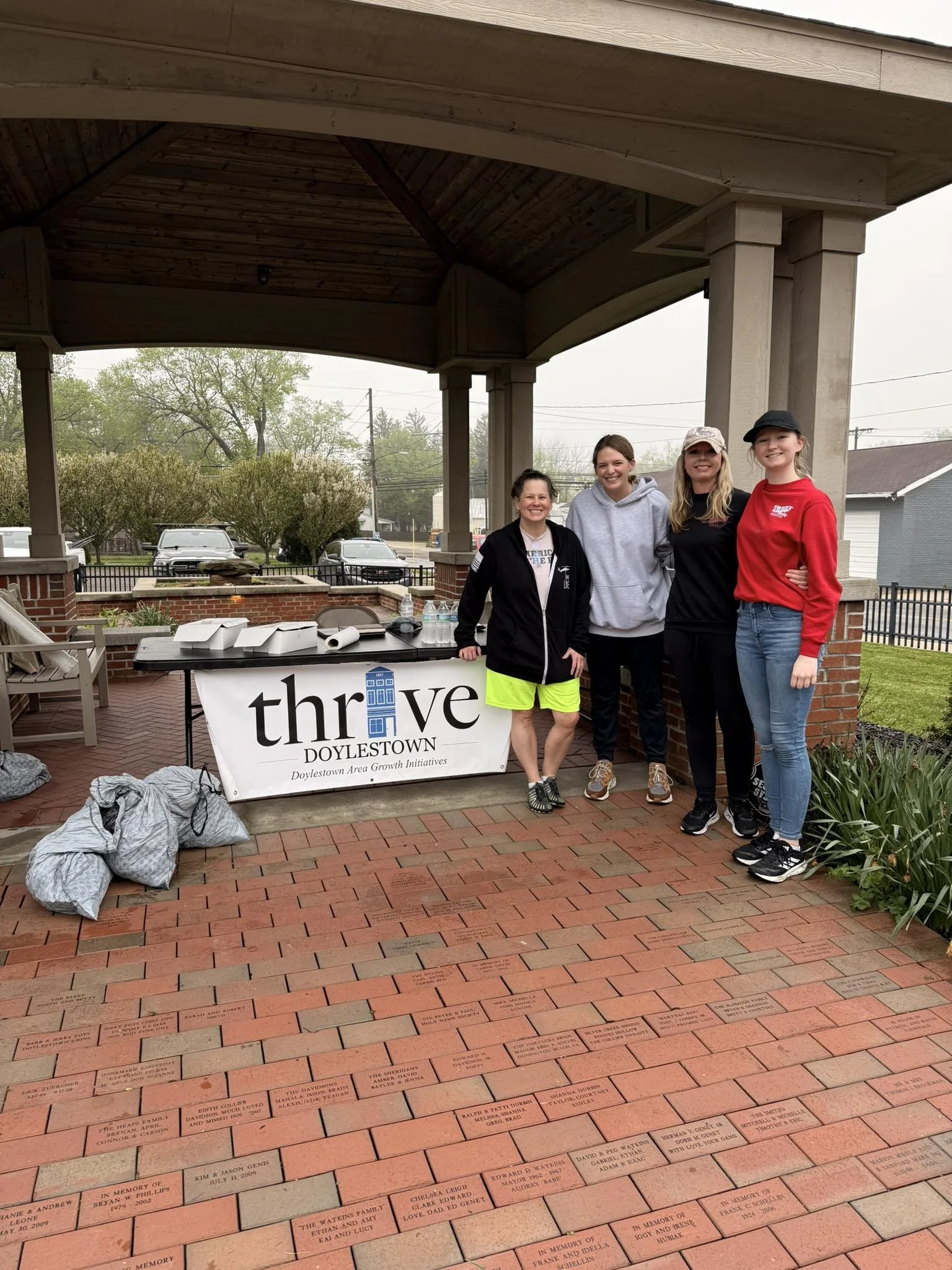 A group of four women and one girl standing together under a large wooden pavilion. In the background, there are trees, cars, and power lines. They are standing behind a table with some water bottles and food containers. There is a banner on the table that says 'Thrive Doylestown' with a blue building icon and the slogan 'Doylestown Area Growth Initiatives.' The ground is paved with bricks engraved with names and memorials.