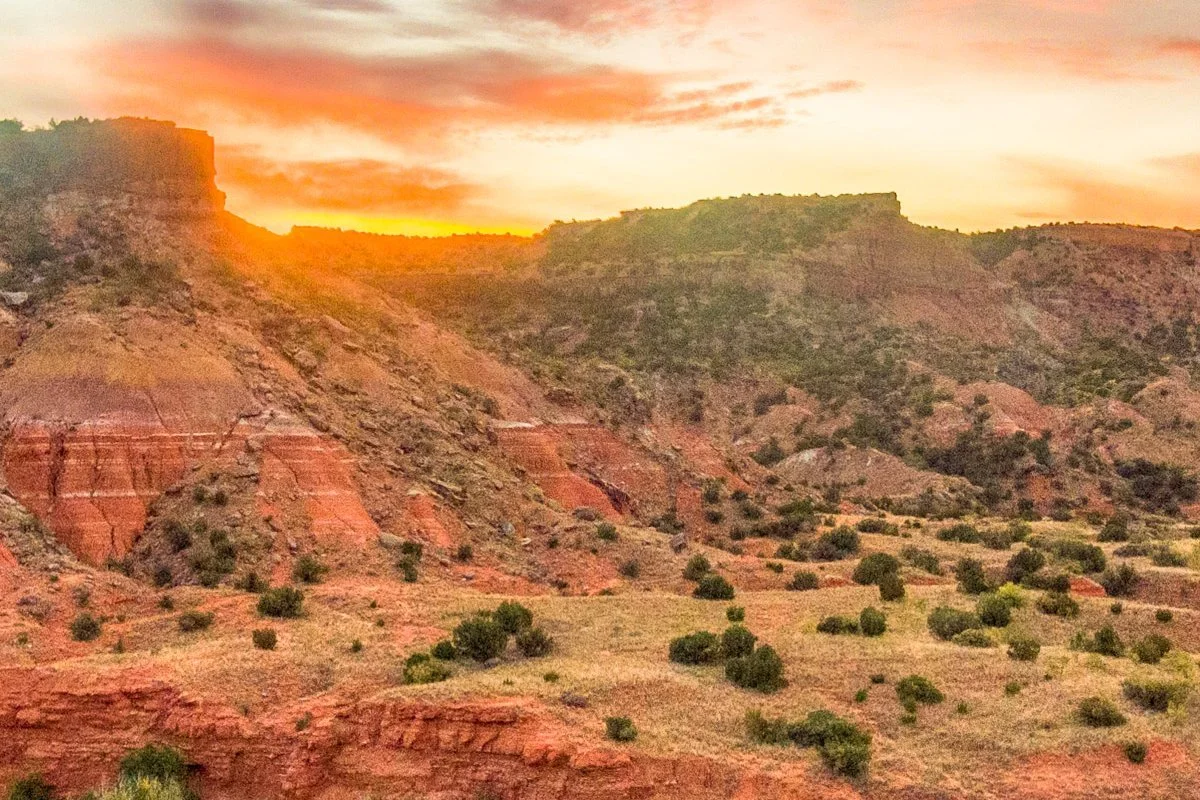 Sunset over a rocky desert landscape with red and brown cliffs and sparse green vegetation.