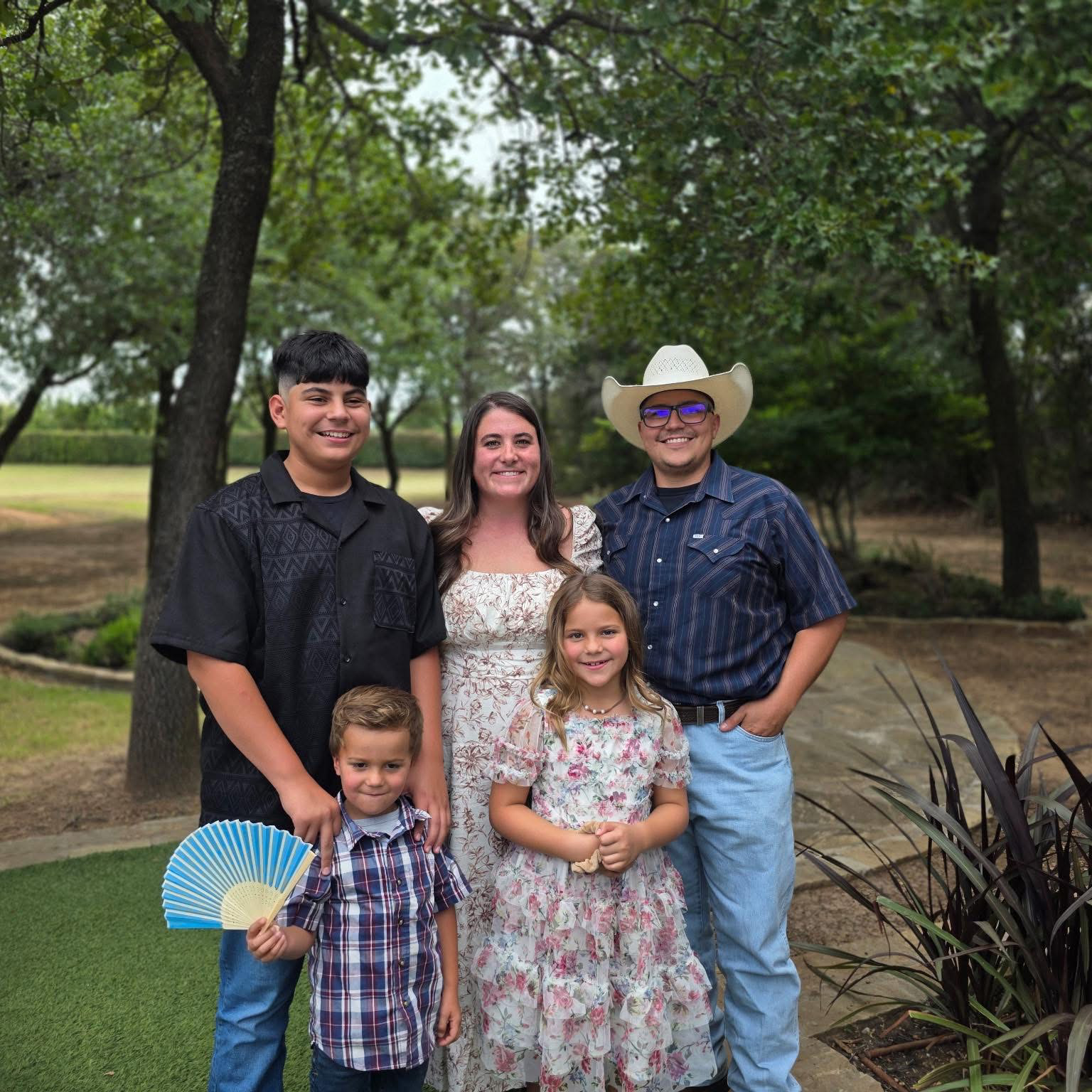 Six people, including children and adults, standing outdoors in a park with trees and greenery, smiling at the camera.
