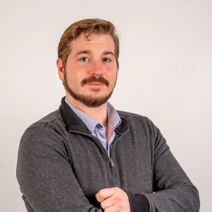 A man with light brown hair, a beard, and mustache in a gray jacket and blue dress shirt, standing against a plain white background, looking at the camera with arms crossed.