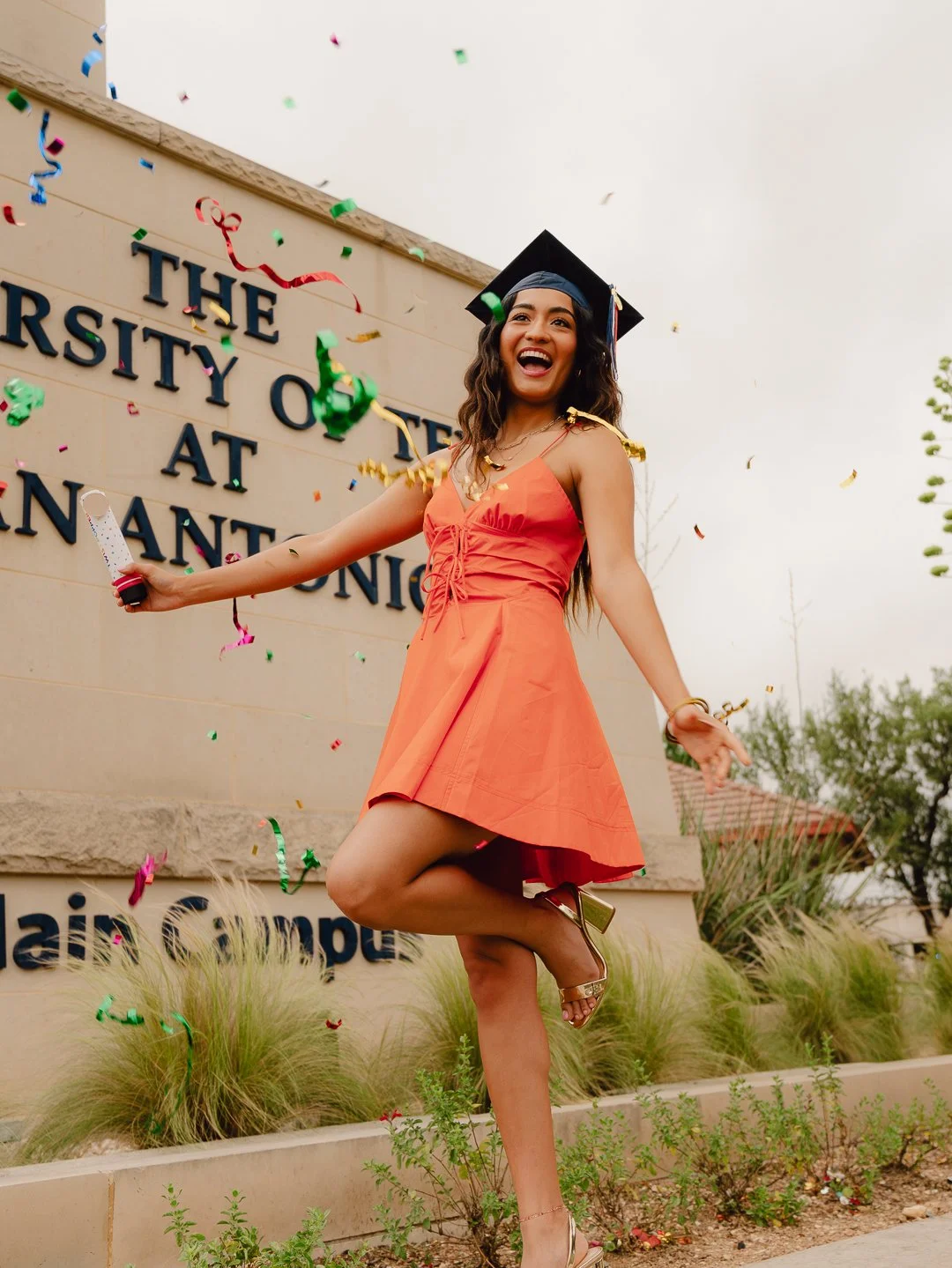 Young woman in orange dress celebrating graduation at university campus, with confetti in the air and a graduation cap on her head.