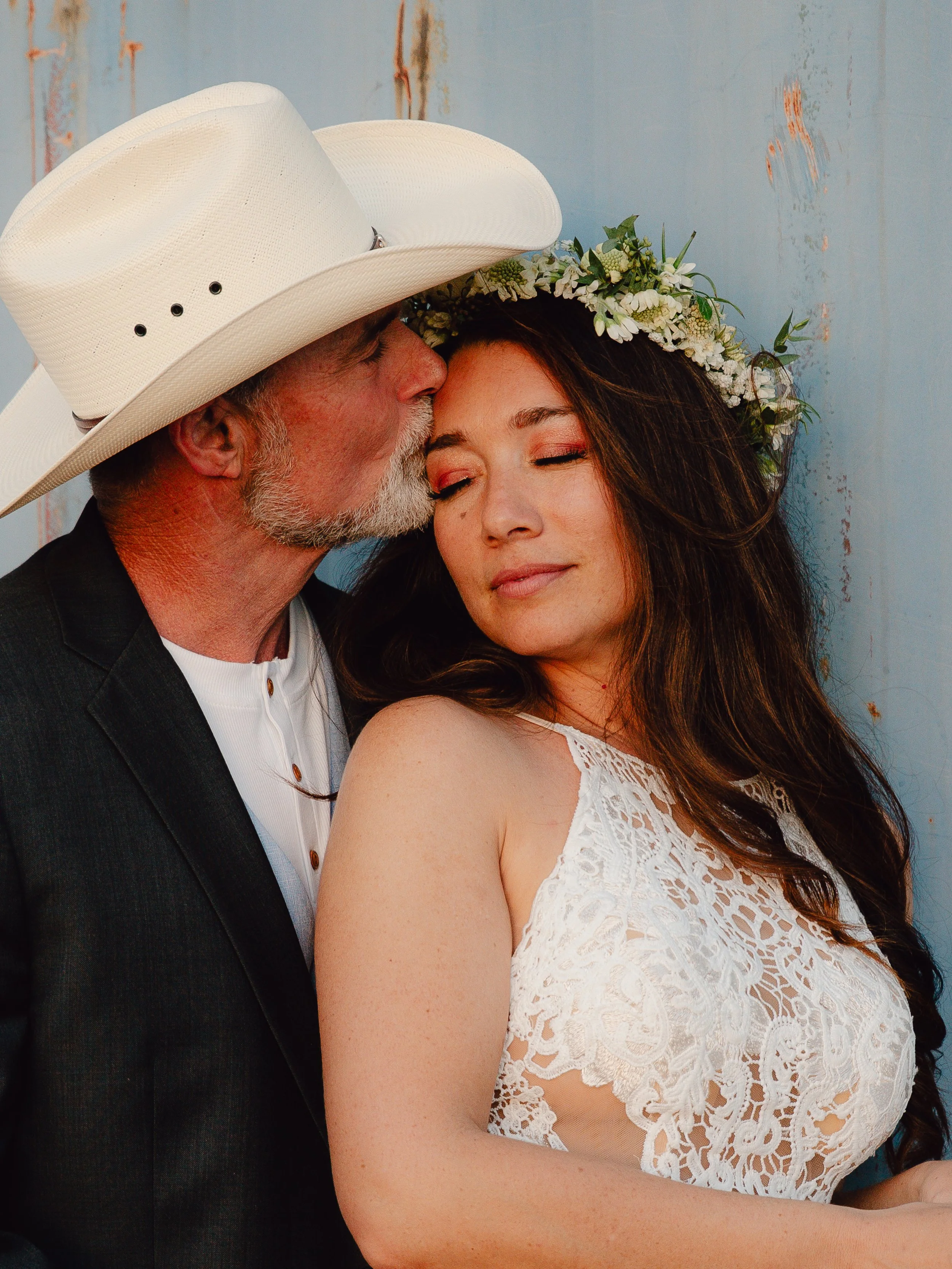 A man with a beard wearing a large white cowboy hat is kissing a woman on the forehead. The woman has long brown hair, closed eyes, and is wearing a white lace dress with a floral crown of white flowers and greenery.