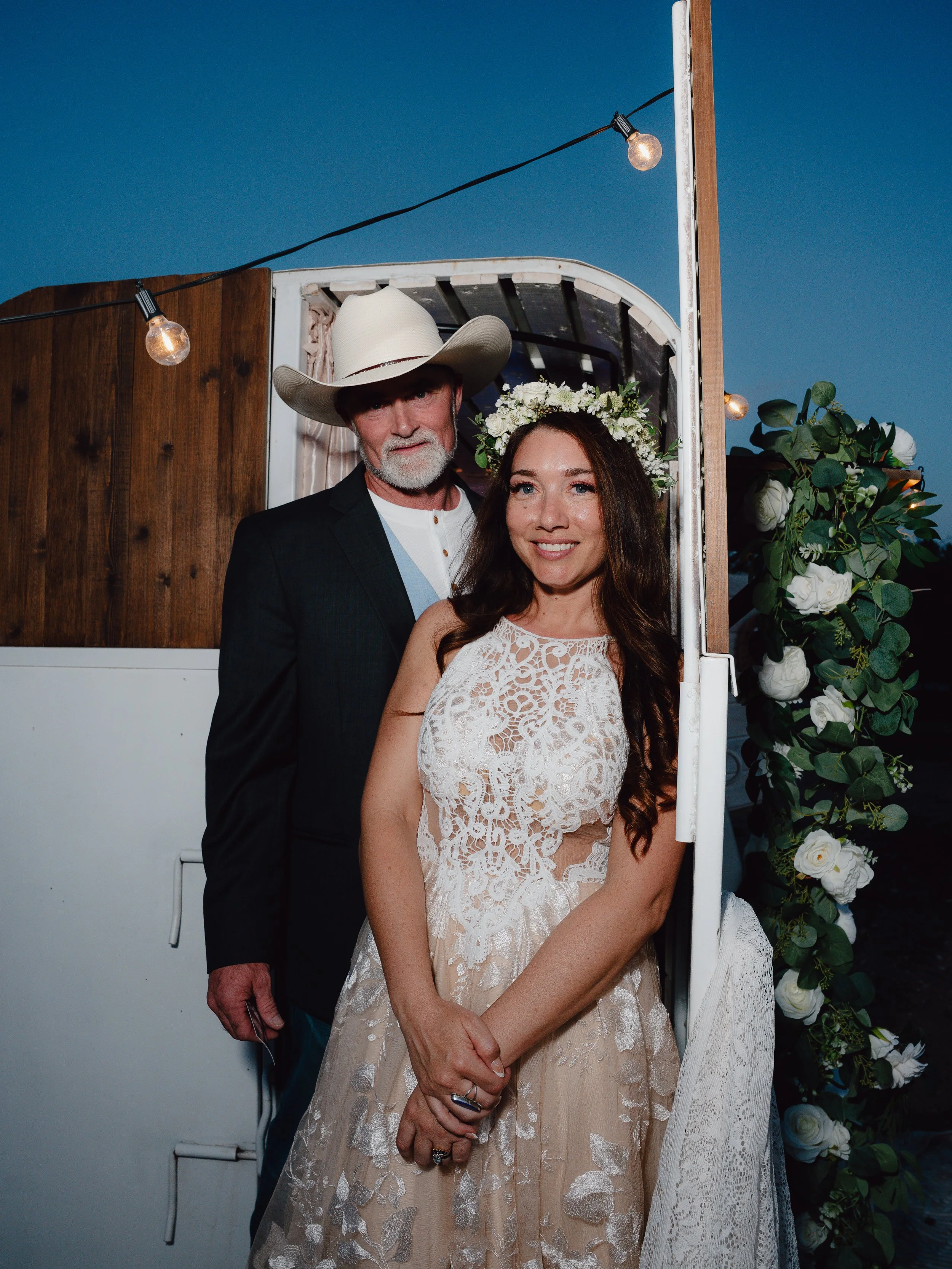 An elderly man and a young woman are posing for a photo at a wedding, with floral decorations and string lights. The man is wearing a white cowboy hat, a dark suit, and a white shirt. The woman is wearing a lace wedding dress and a floral crown with 
