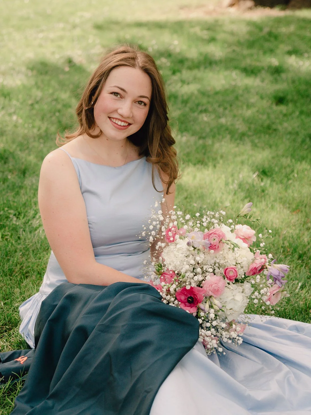 A young woman with brown hair, wearing a sleeveless light blue dress, sitting on grass outdoors and holding a large bouquet of pink, white, and lavender flowers.
