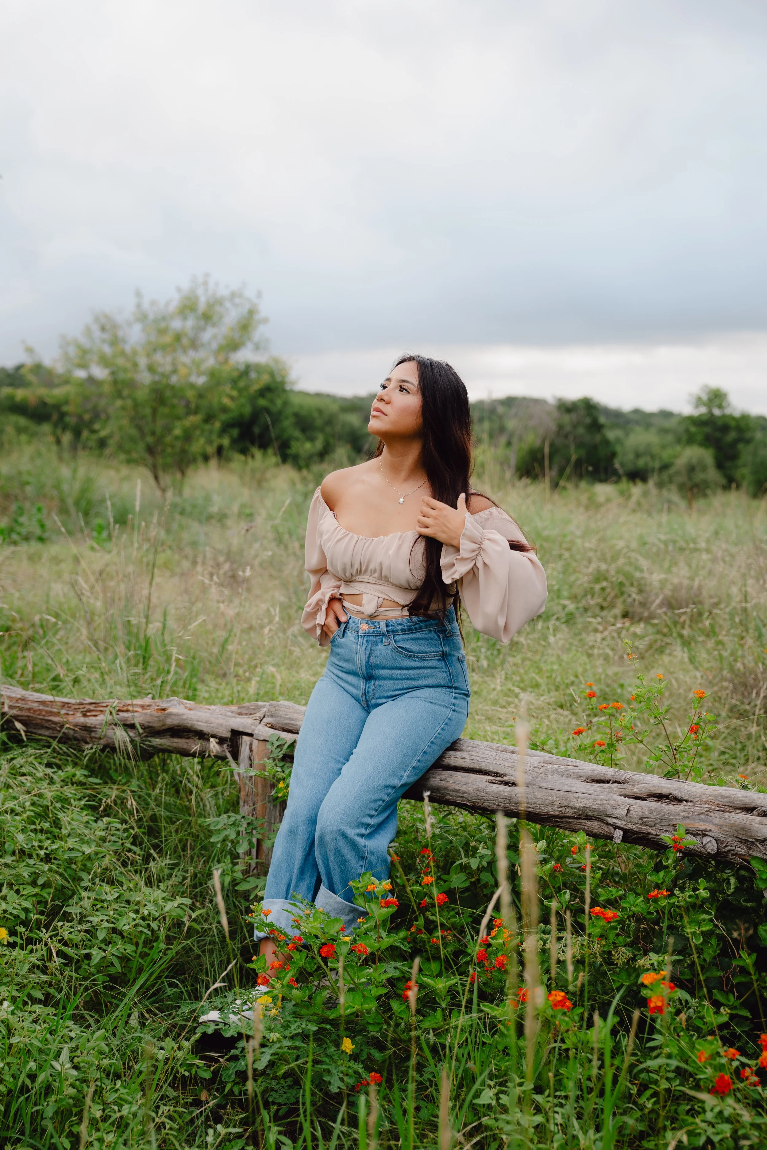 A woman wearing a beige off-the-shoulder top and high-waisted blue jeans sitting on a log in a grassy field with flowers, looking up at the sky under a cloudy sky.