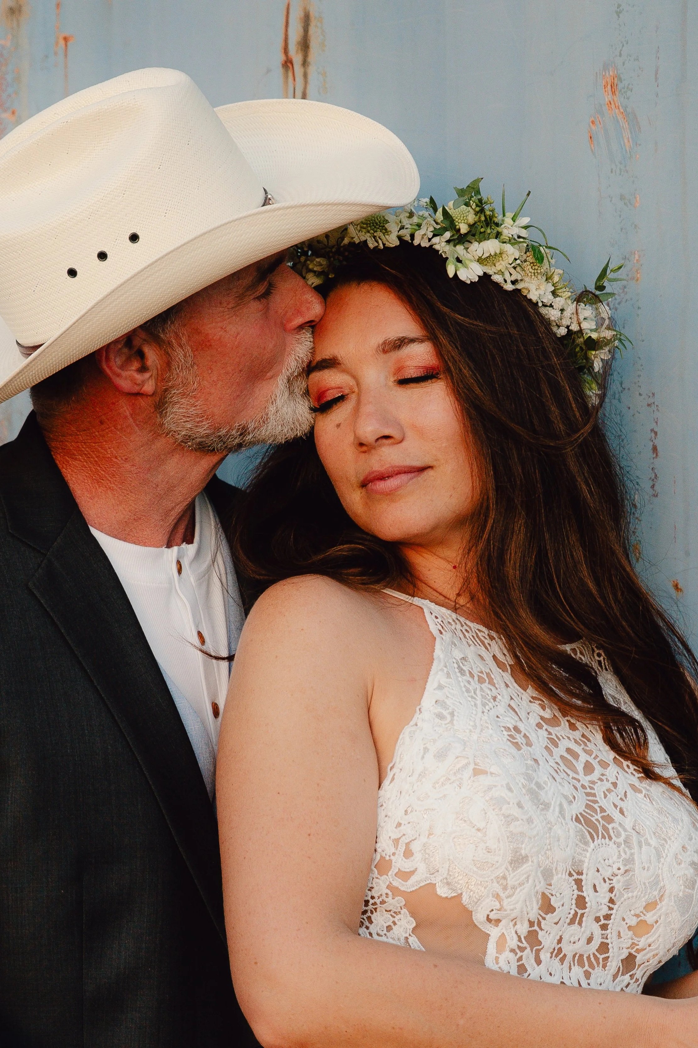 An older man with a gray beard wearing a white cowboy hat, kissing a young woman on her forehead. The woman has long brown hair, wearing a floral crown and a white lace top, with a peaceful expression and closed eyes.