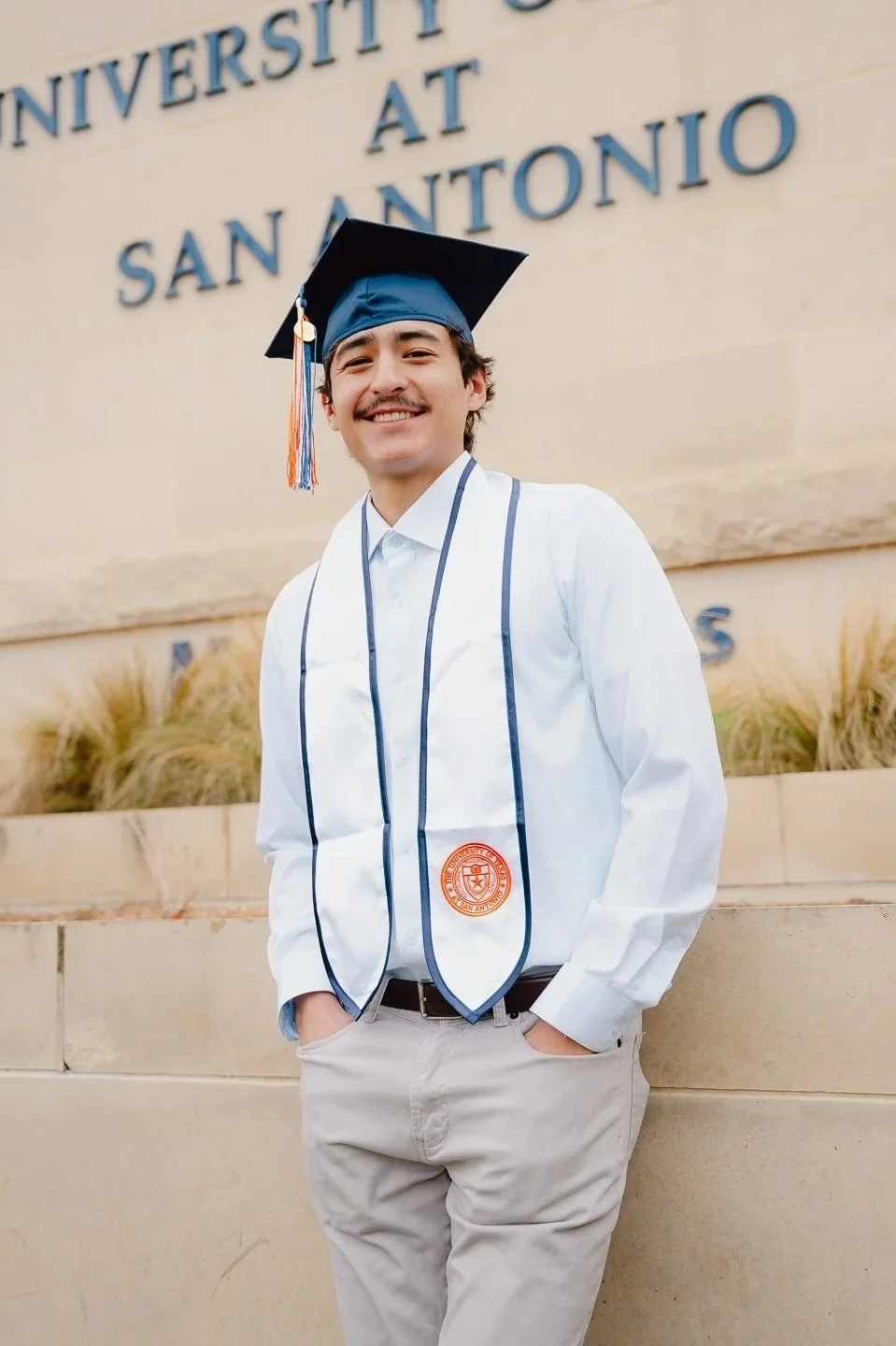 A young man smiling in graduation cap and gown standing outside in front of a university sign at San Antonio.