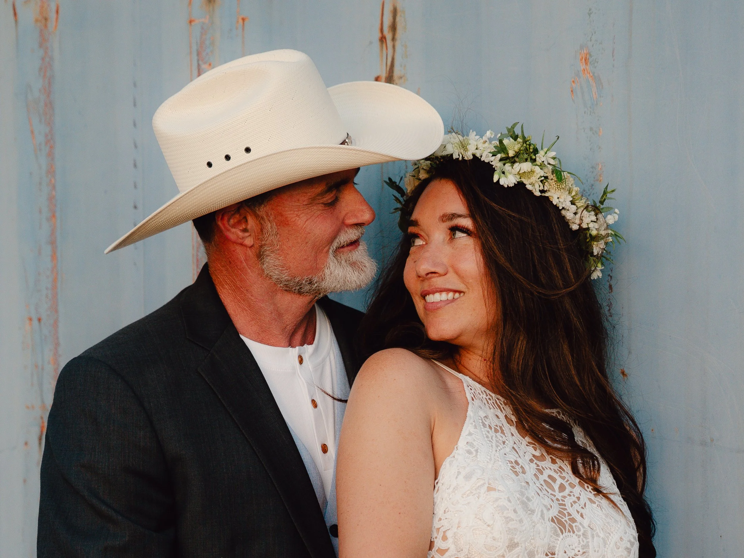 A man and a woman face each other close-up, with the man wearing a white cowboy hat and the woman wearing a floral headband. The background is a rustic, weathered blue wall.