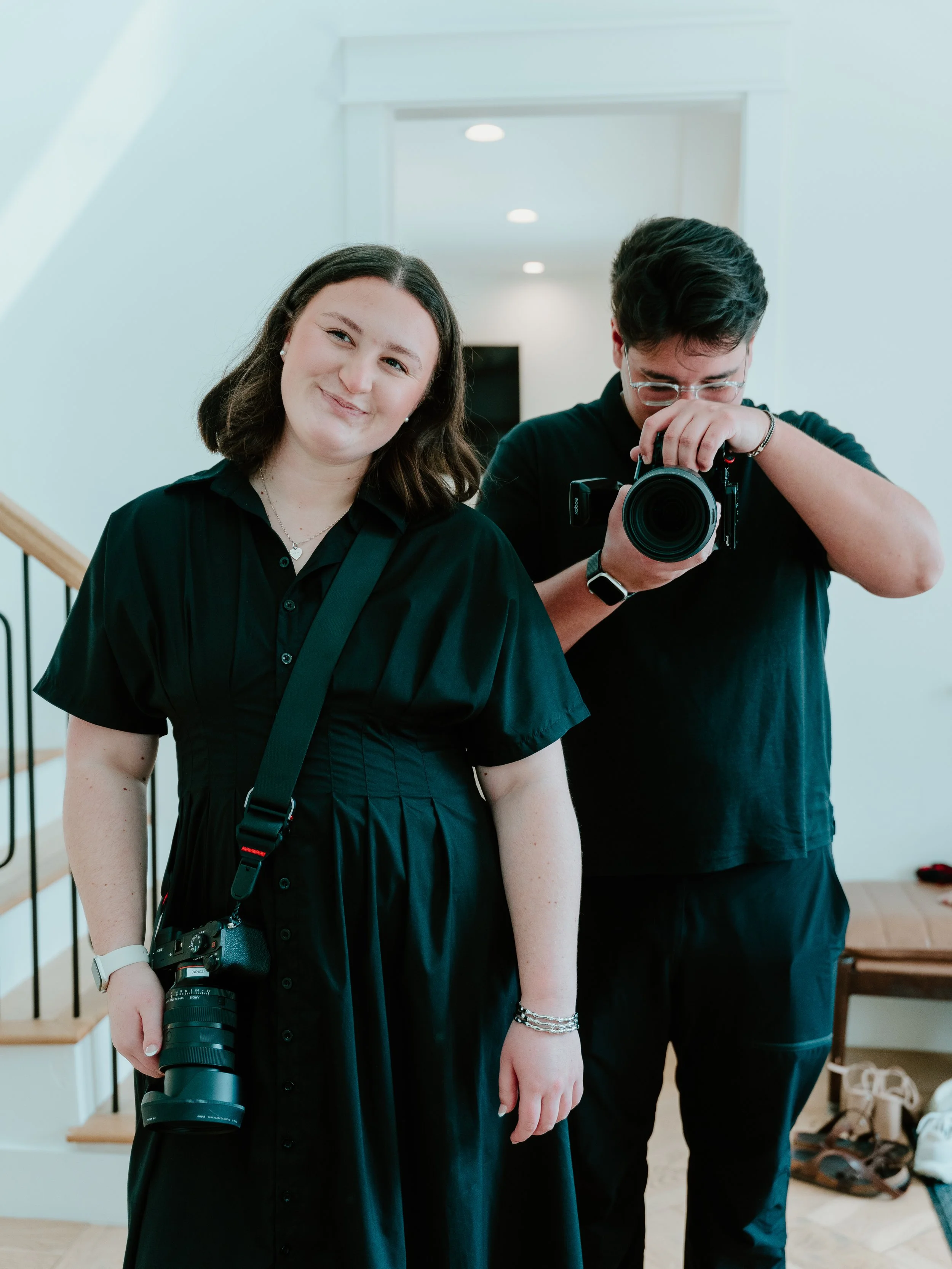 Two young adults, a woman and a man, taking a mirror selfie in a bright indoor space. The woman is smiling slightly, wearing a black dress, with a camera hanging from her neck. The man is looking down while adjusting his glasses, holding a professional camera with a long lens. Shoes and bags are visible on the floor in the background.