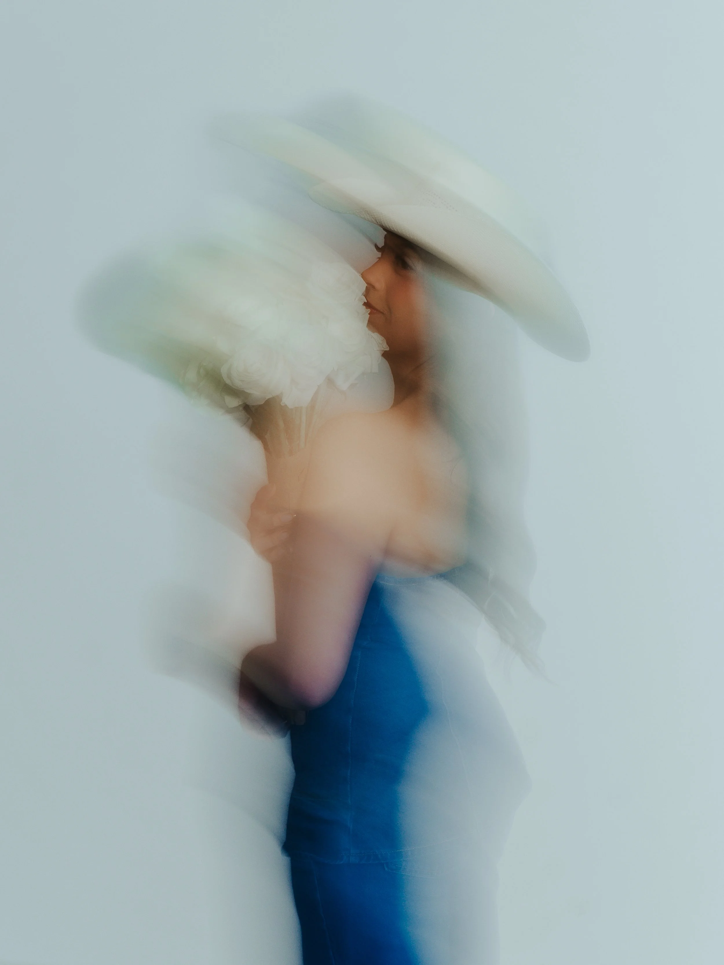 A person standing against a plain white wall, wearing a large floppy hat and a blue dress, holding a white flower.