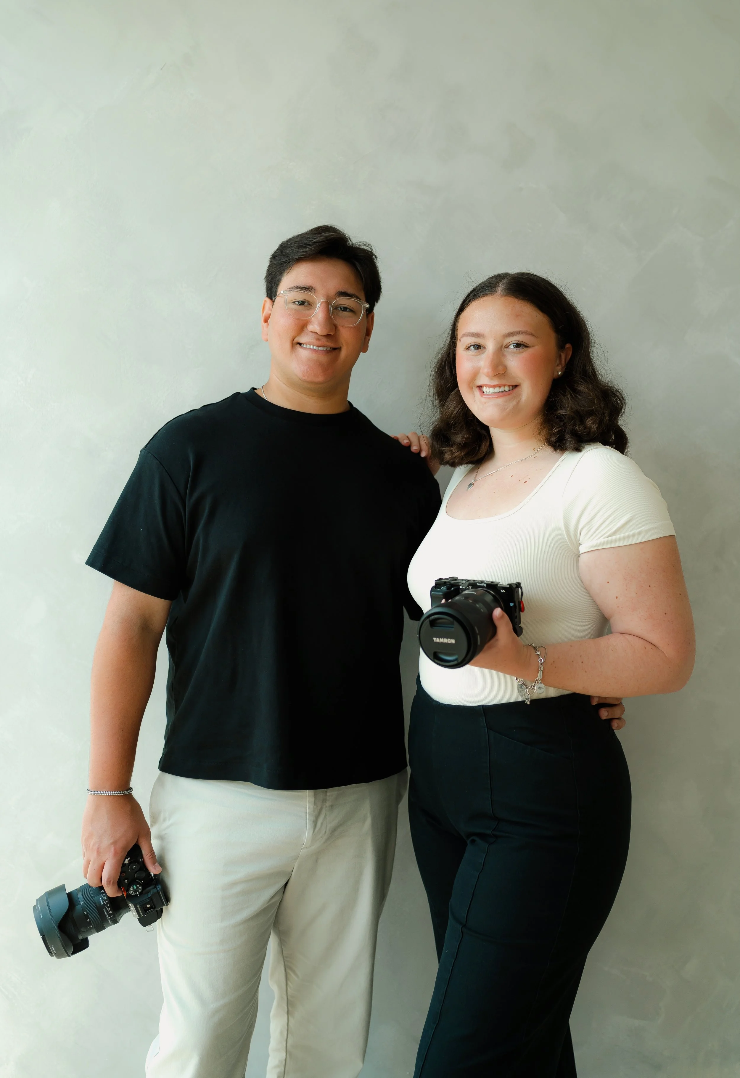 A man and a woman standing together in front of a plain wall, both smiling. The man is holding a camera with a large lens and the woman is holding a camera. They appear to be photographers or photography enthusiasts.