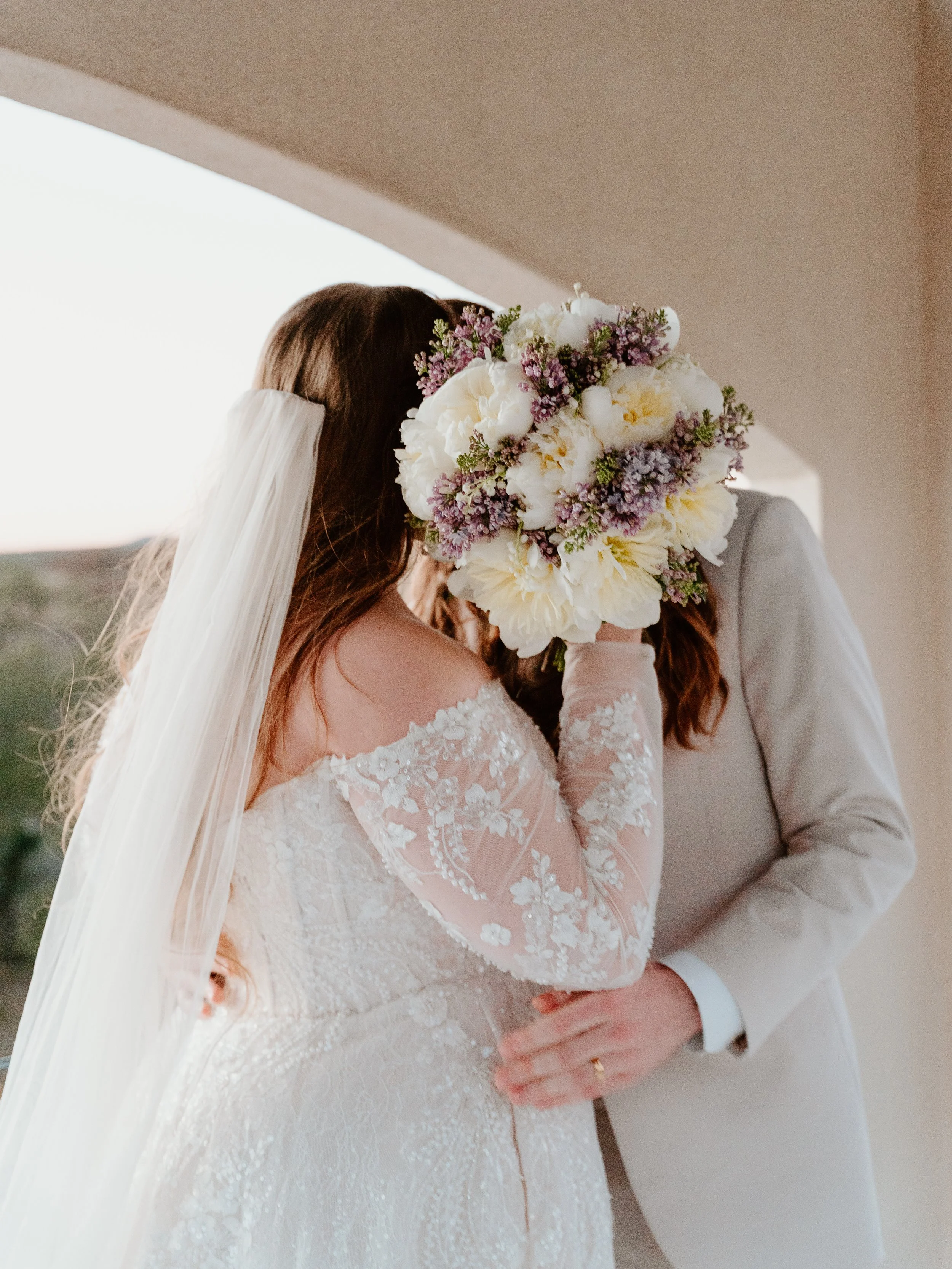 A bride in a white lace wedding dress and veil holding a bouquet of white and purple flowers, standing close to a groom in a beige suit and white shirt.