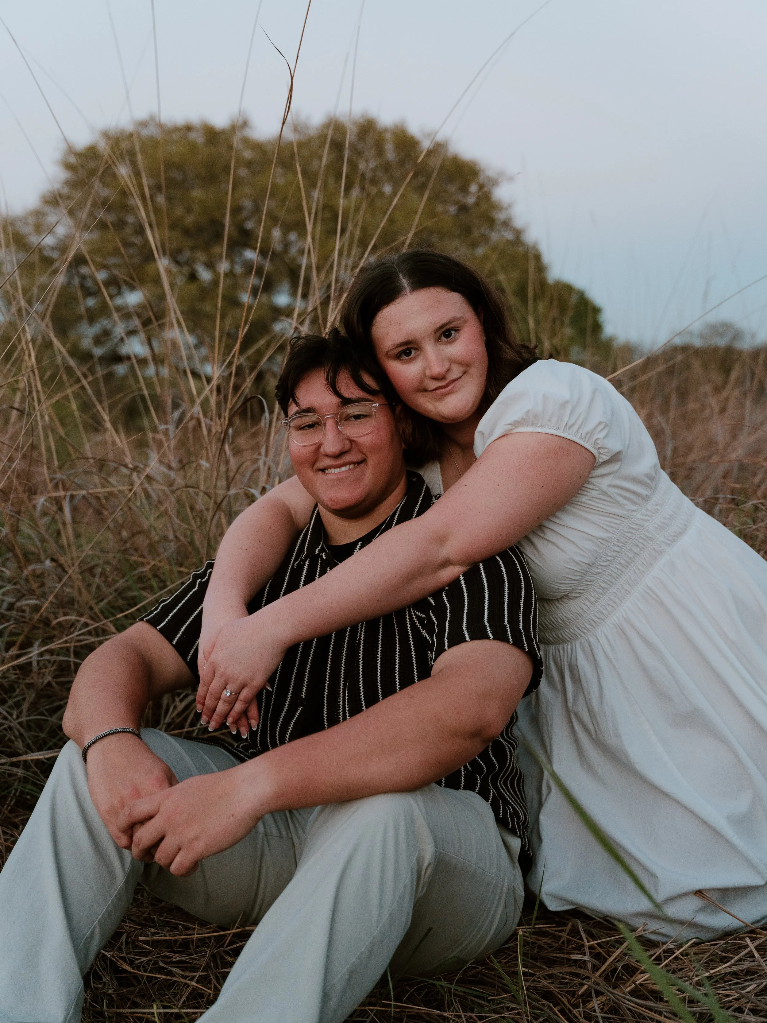 Two young women sitting among tall grass in an outdoor setting, embracing each other, smiling at the camera, with trees in the background and dusk light.