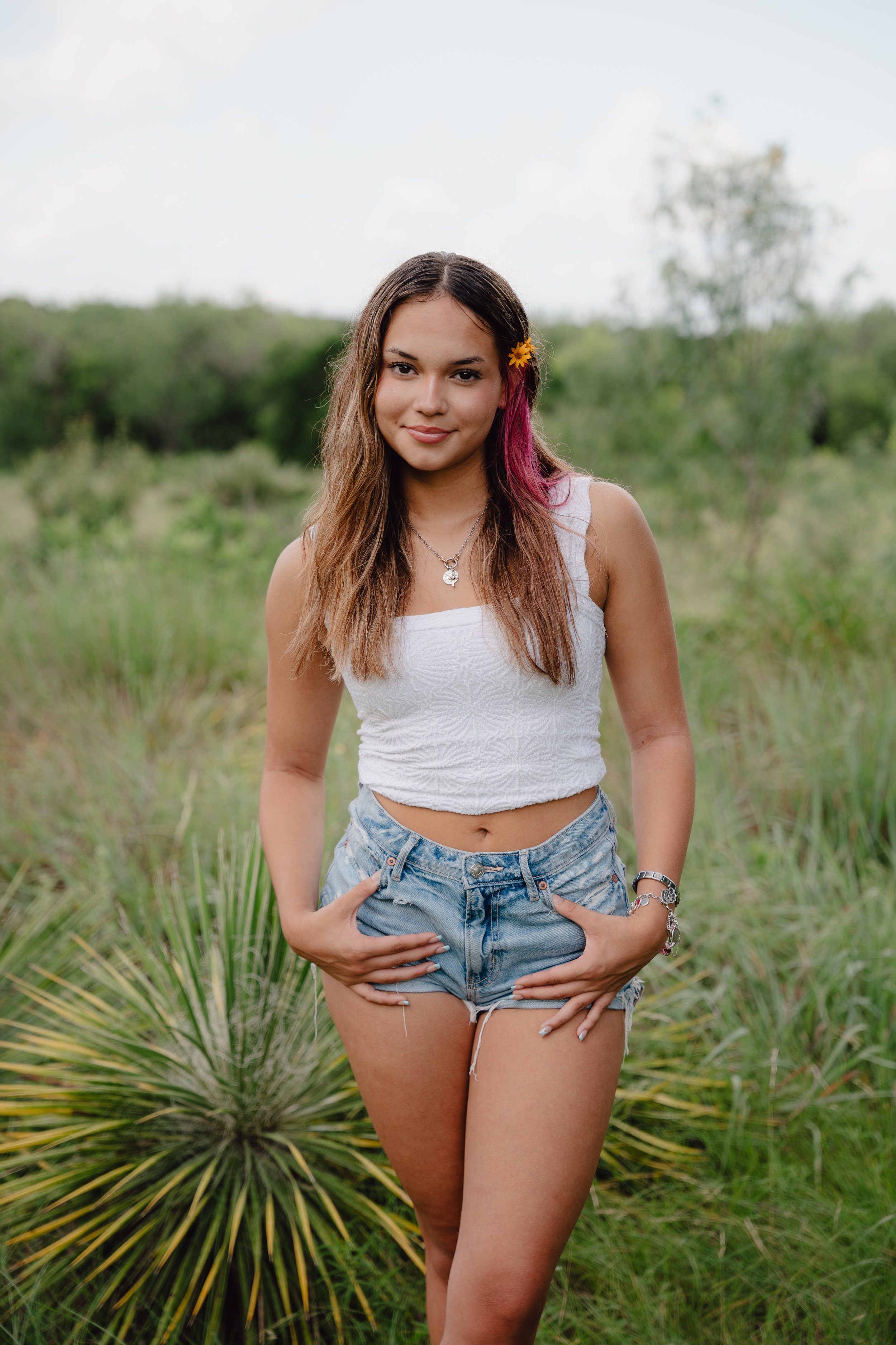 Young woman with long brown hair, pink highlights, and a yellow flower in her hair, standing outdoors in a green field with bushes and trees in the background. She is wearing a white sleeveless top, denim shorts, and various jewelry, and has her hands in her pockets.
