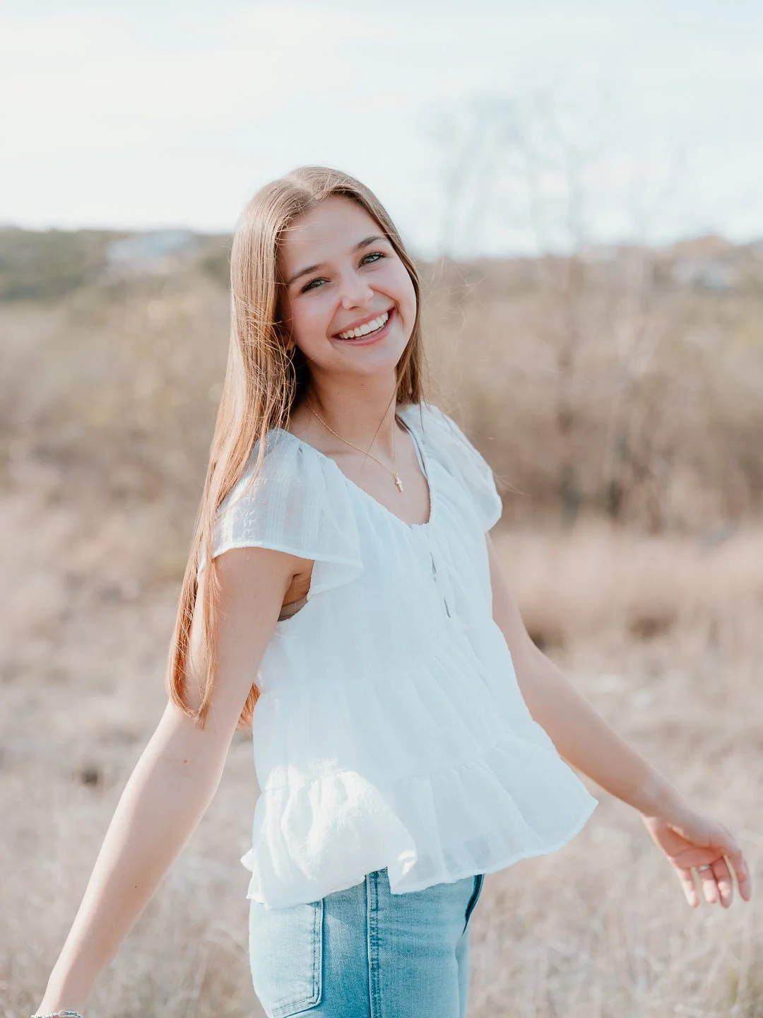 Young woman with long light brown hair smiling outdoors in a field, wearing a white blouse and blue jeans.
