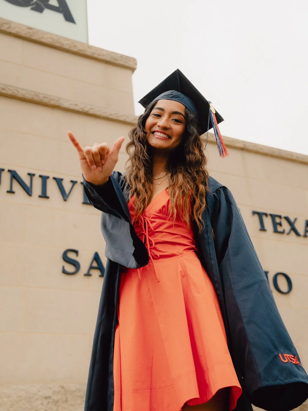 A smiling woman in a graduation cap and gown standing in front of a sign that reads 'University of Texas at San Antonio' making a shaka hand gesture.