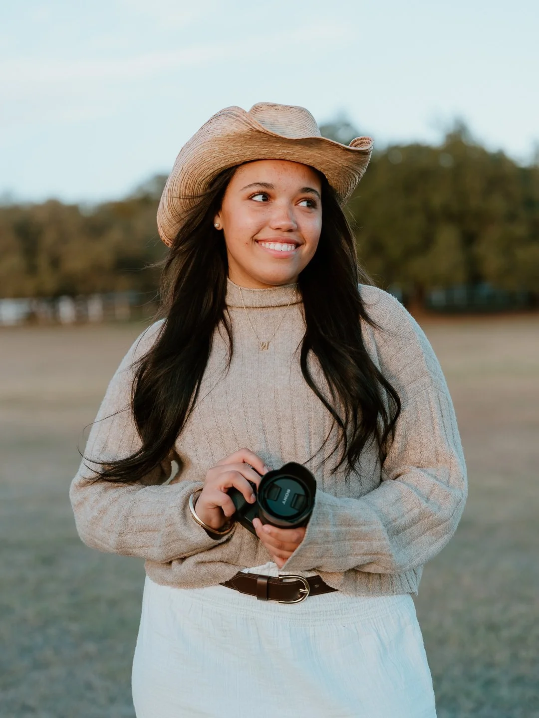Young woman wearing a beige turtleneck sweater, a brown belt, and a wide-brimmed hat, holding a camera in an outdoor park setting with trees in the background.