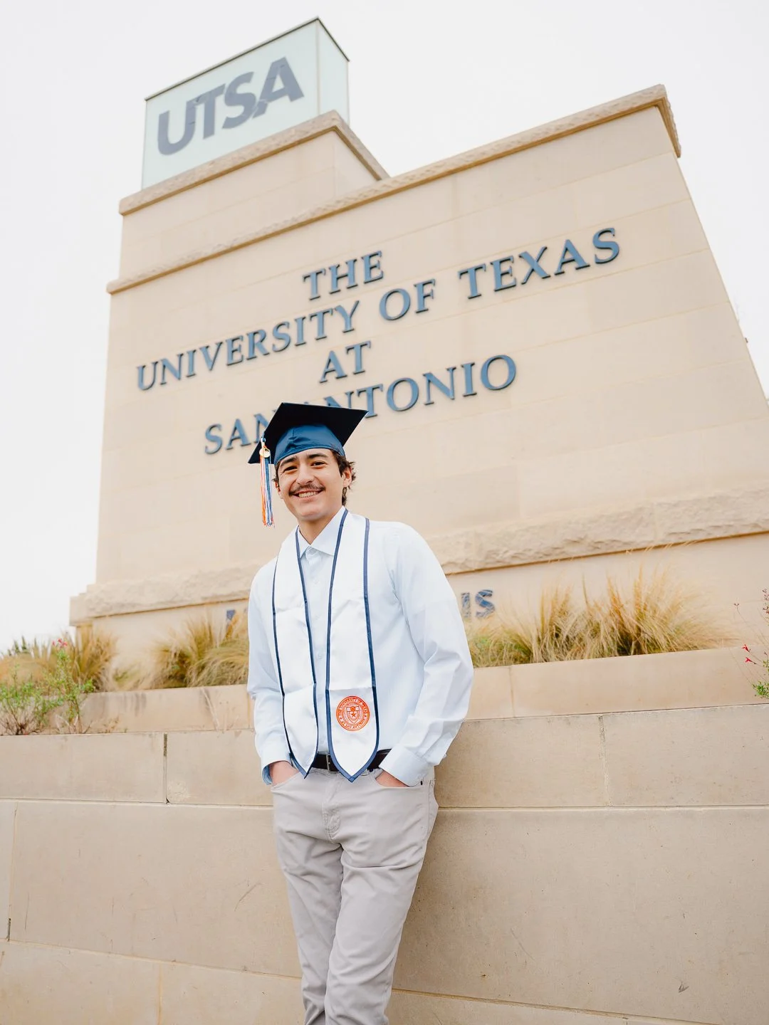 A smiling young man in graduation attire, including a cap and gown with a cord, standing in front of a large university sign that reads "The University of Texas at San Antonio."