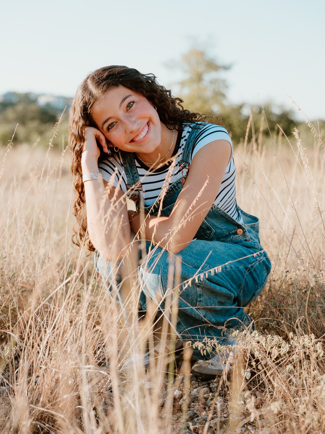 Young woman with curly hair smiling, sitting in a field of tall grass during daytime.